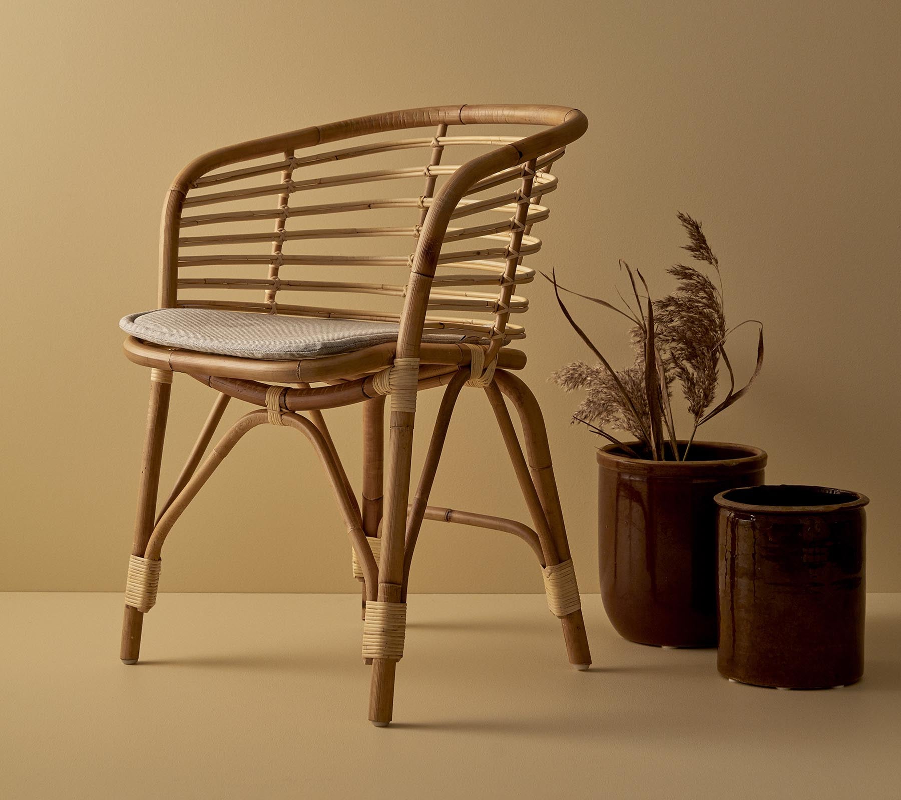 En stilfuld rattan stol med polstret sæde, ledsaget af to dekorative krukker med tørrede planter.
