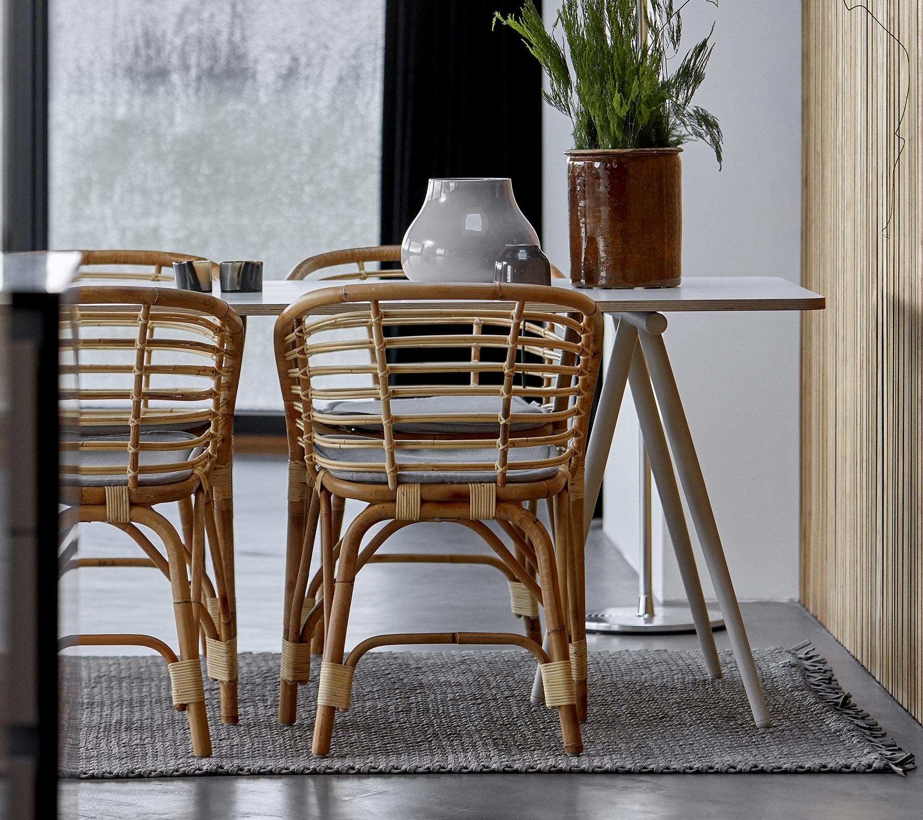 Et moderne spisearrangement med rattanstole, et slankt bord og dekorativt grønt i et stilfuldt interiør.