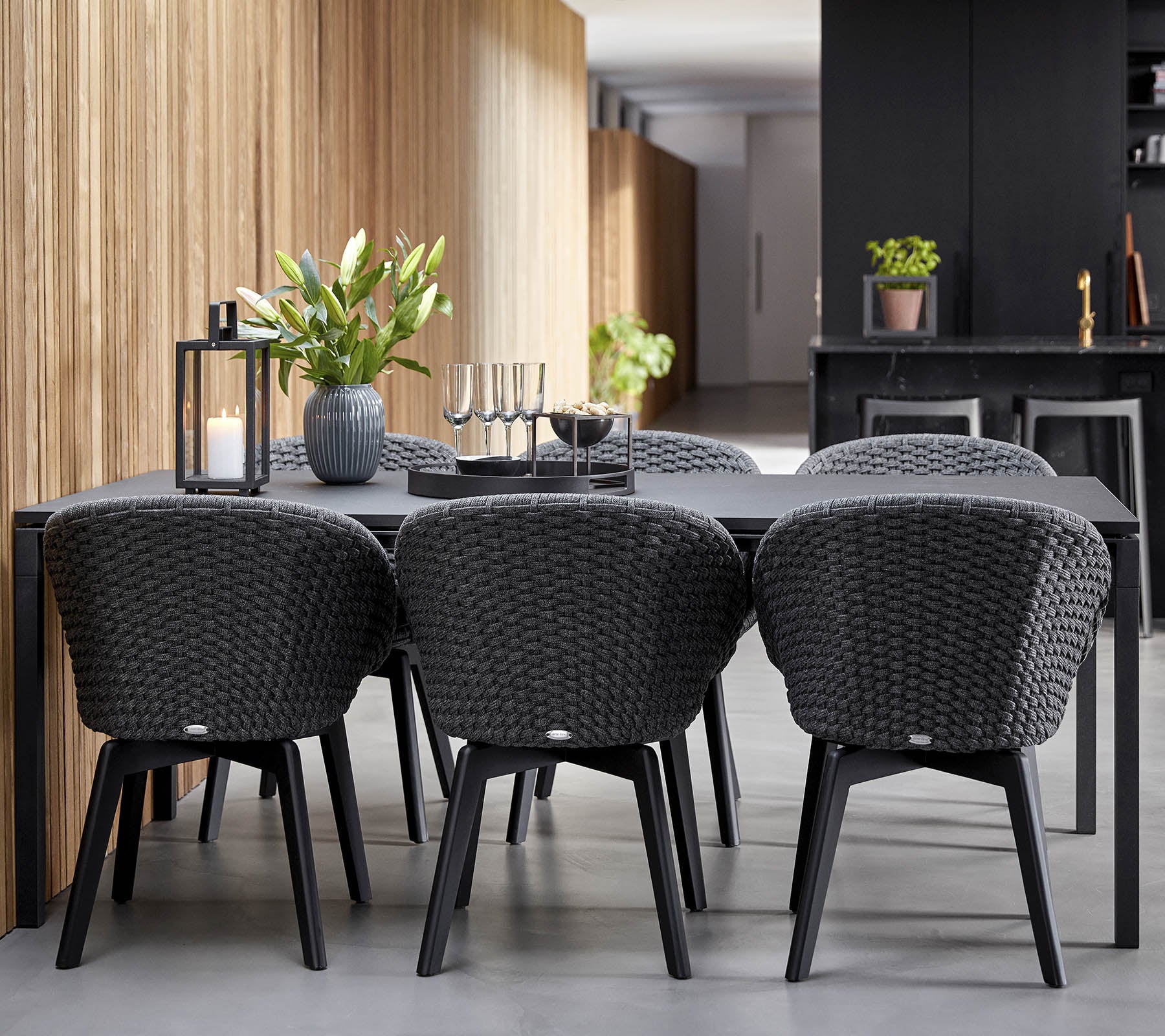 Et moderne spisestueopstilling med et slankt sort bord omringet af stilfulde vævede stole, suppleret af dekorative planter og lys.