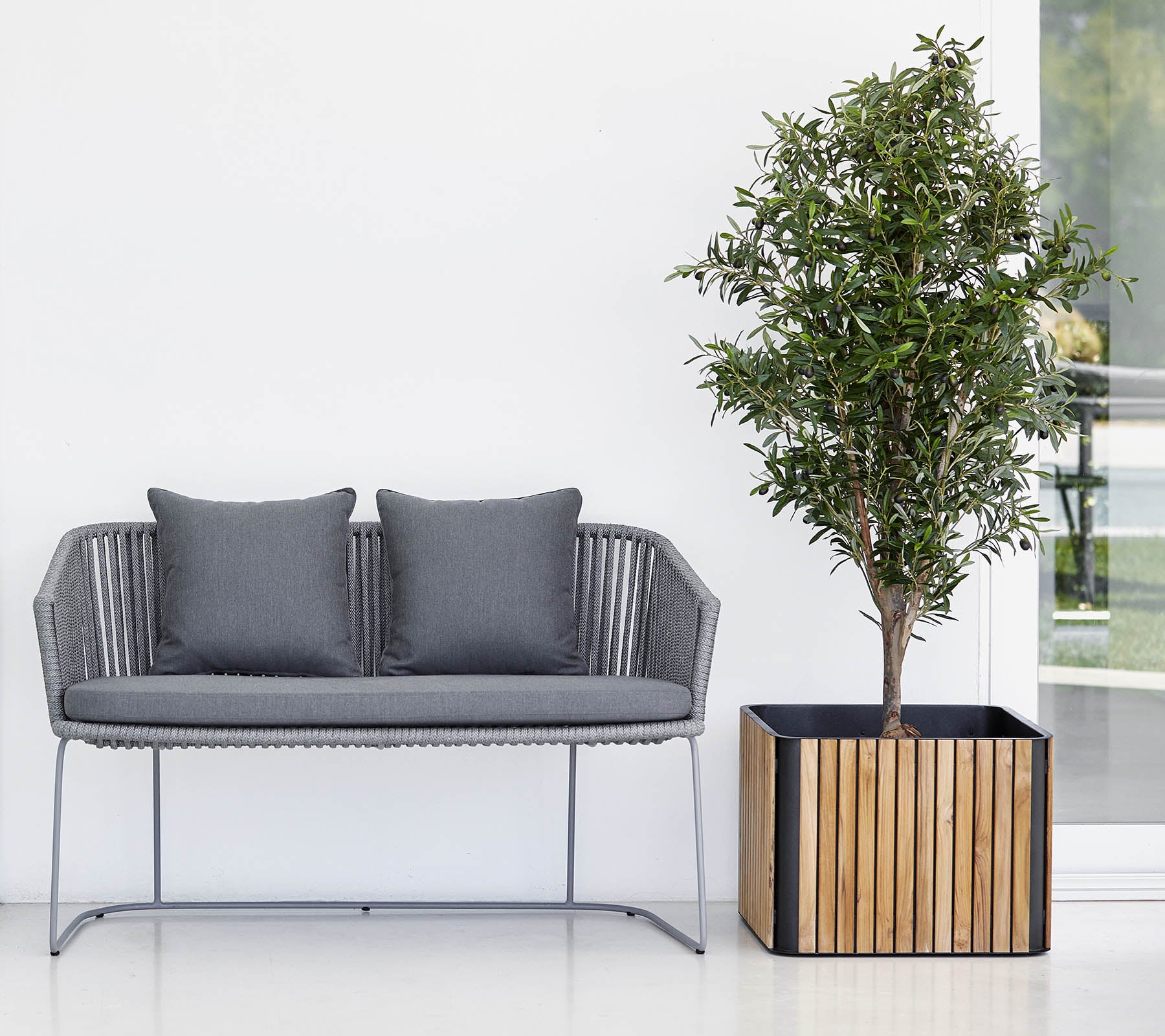 Moderne sofa med to puder ved siden af et potteplante i en dekorativ krukke, mod en ren, neutral baggrund.