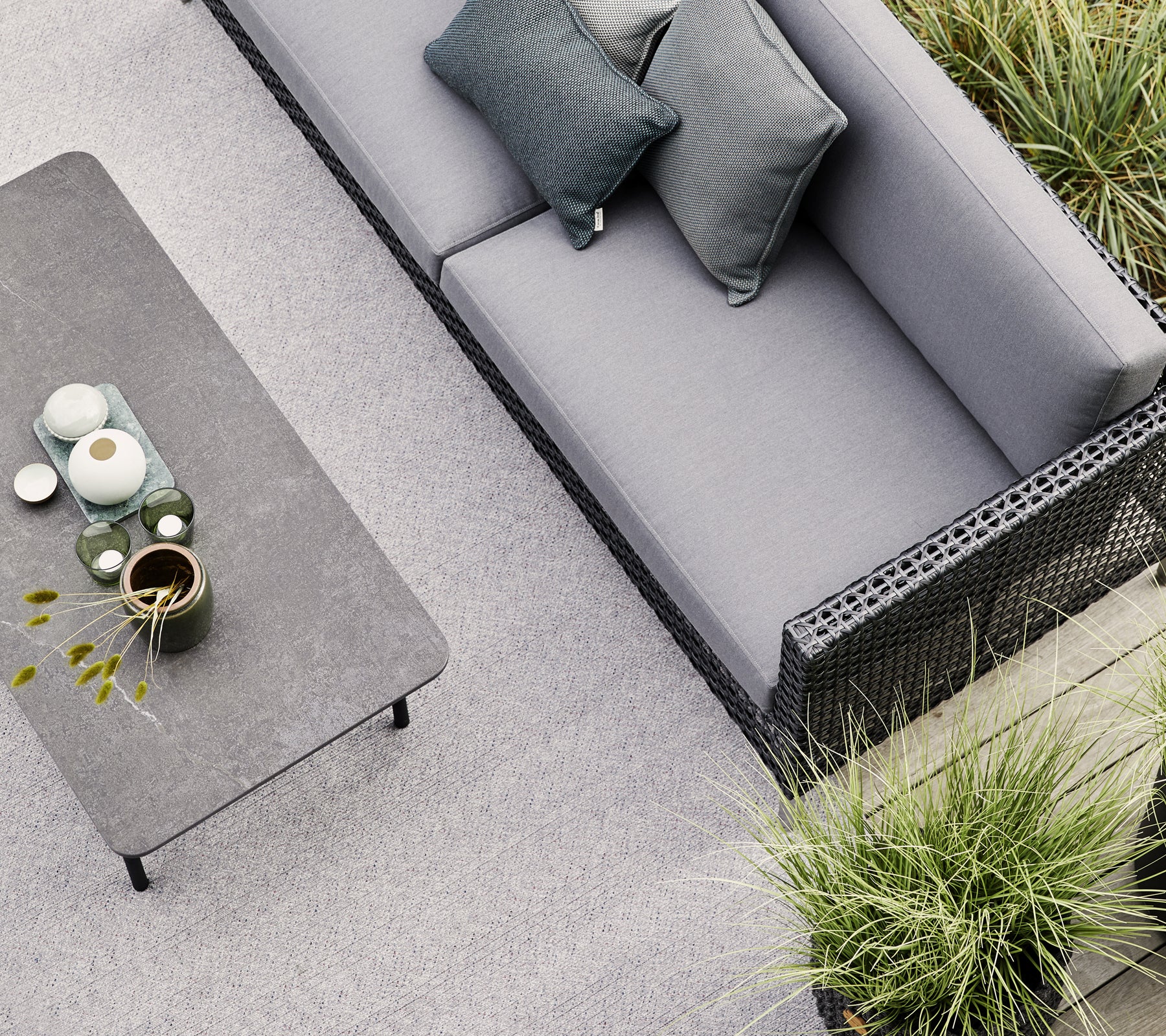 En grå sofa med to puder står nær et minimalistisk bord prydet med keramik og en lille plante, omgivet af grønne planter.