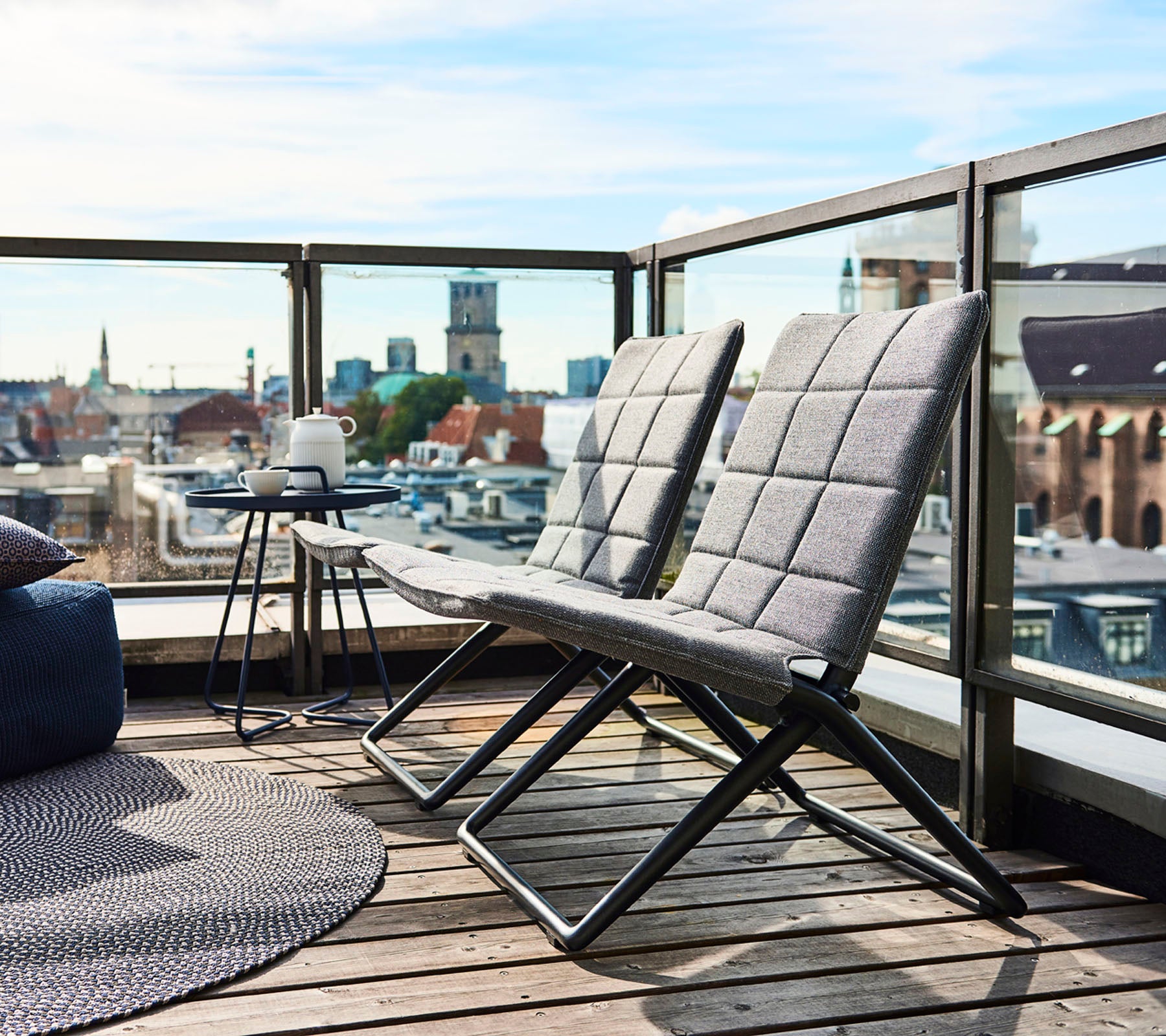 To moderne loungestole med slanke rammer og teksturerede puder, ledsaget af et lille sidebord og udsigt over byen i baggrunden.