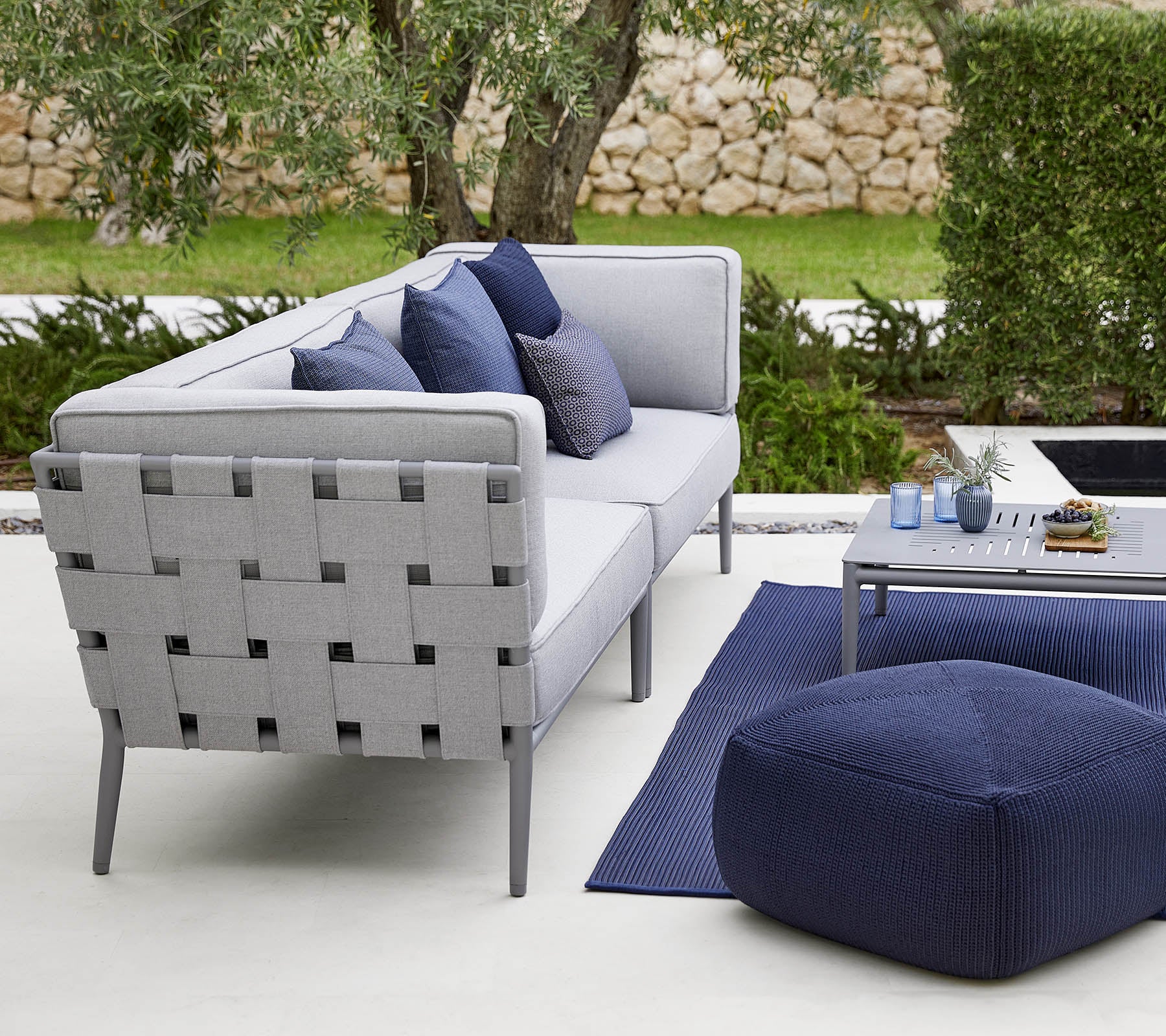Moderne udendørs siddearrangement med en grå sofa med dekorative puder, et stilfuldt kaffebord og en hyggelig pouf.