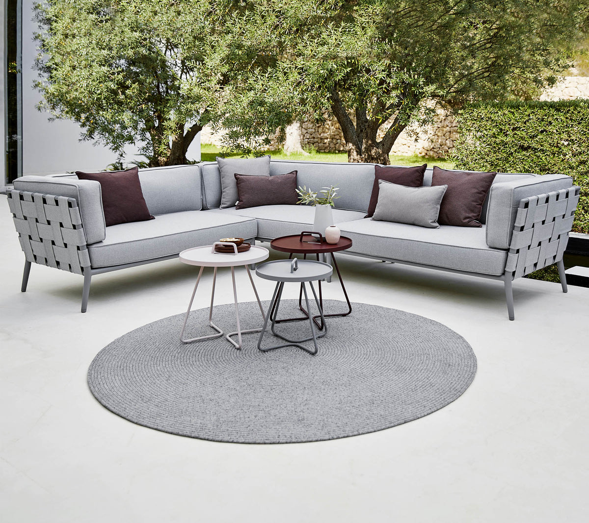 Moderne sectional sofa i lys grå med dekorative puder, ledsaget af stilfulde sofaborde på et rundt tæppe. Luksuriøs grønne planter i baggrunden.