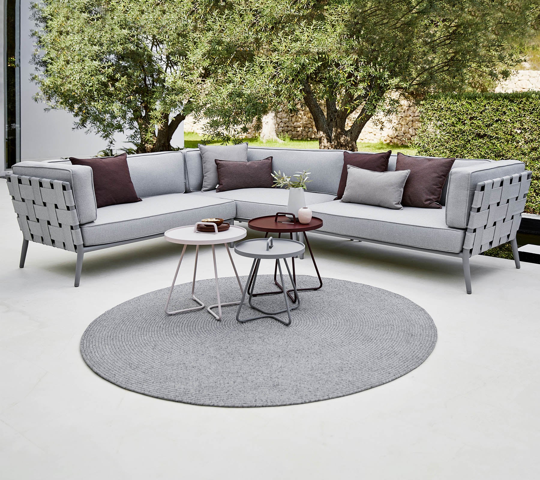 Moderne sectional sofa i lys grå med dekorative puder, ledsaget af stilfulde sofaborde på et rundt tæppe. Luksuriøs grønne planter i baggrunden.