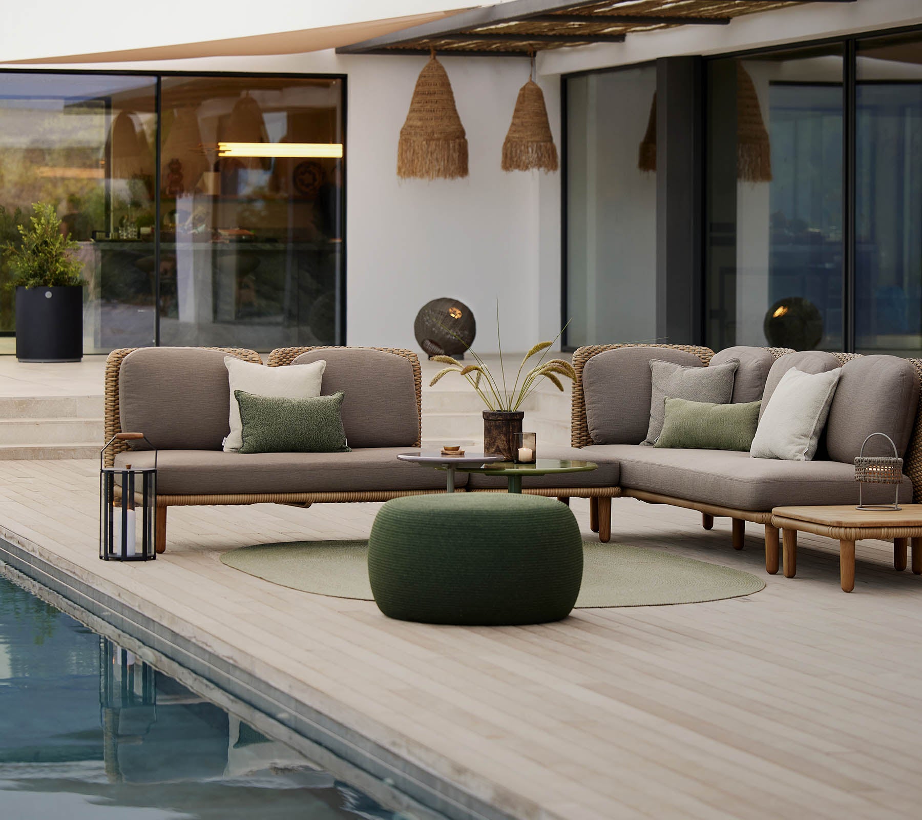 Et stilfuldt loungeområde med en sektionsofa med puder, et rundt bord og en hyggelig pouf, der ligger ved siden af en rolig pool.