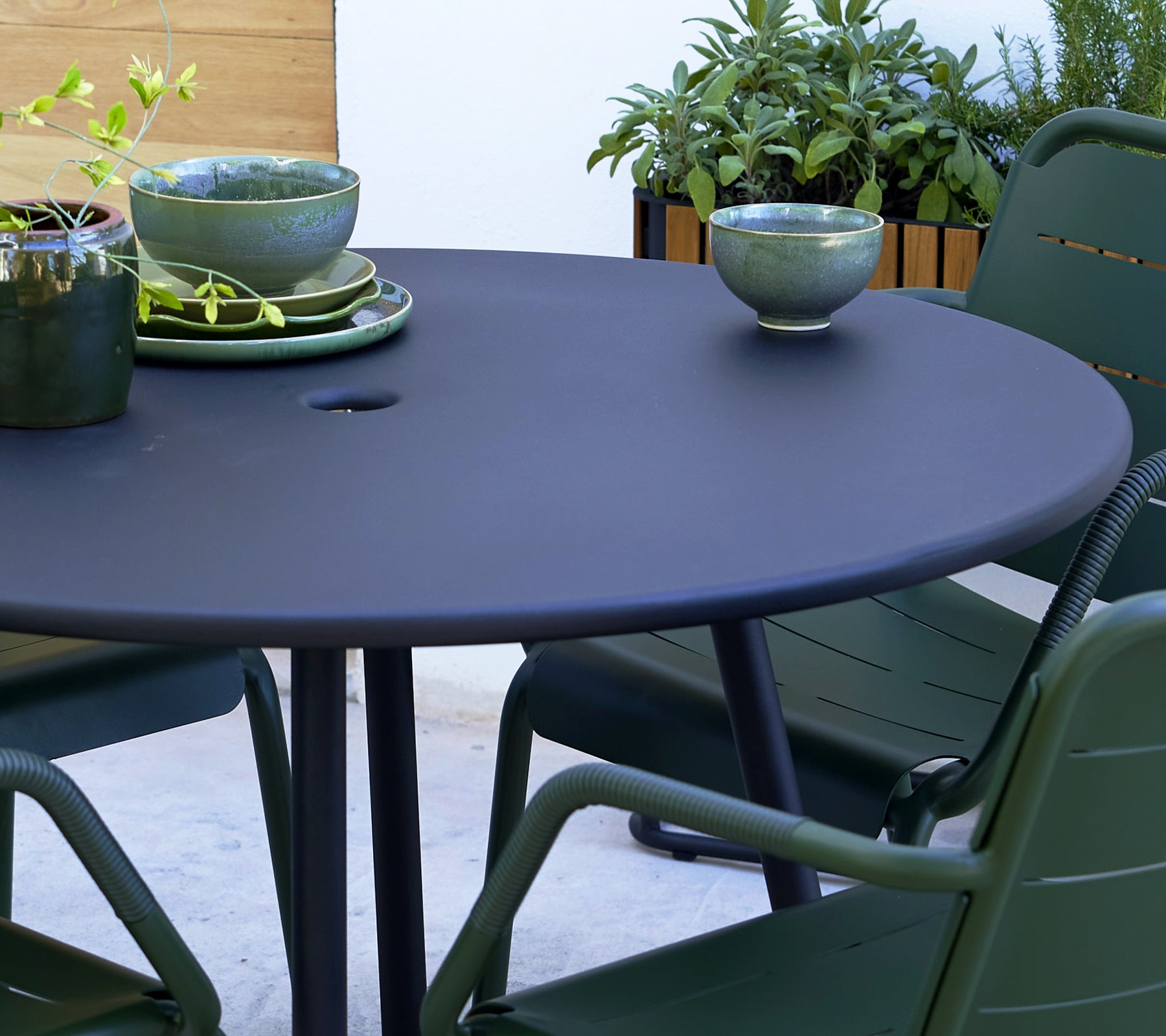 Rund grå tabel med grønne København-stole, prydet med dekorative skåle og planter i baggrunden.