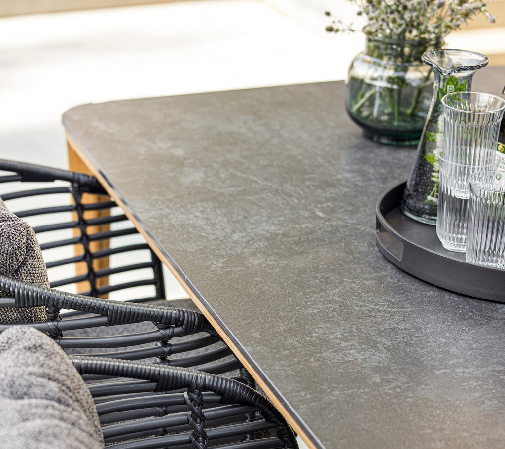 Et moderne spiseopstilling med en glat bordplade, stilfuld kurvestol, glasvarer på en bakke og en dekorativ vase med grønne planter.
