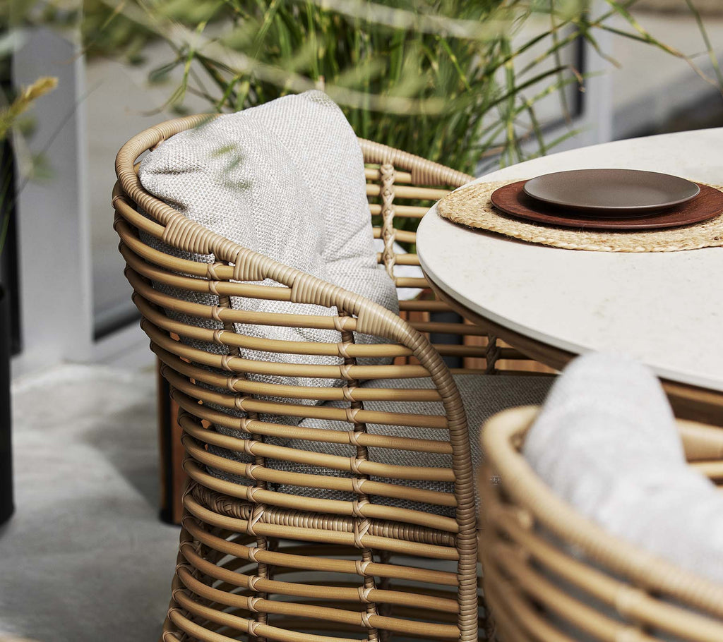 Basket chair with a curved design, featuring comfortable cushions and a nearby table set with a plate and placemat. Greenery in the background.