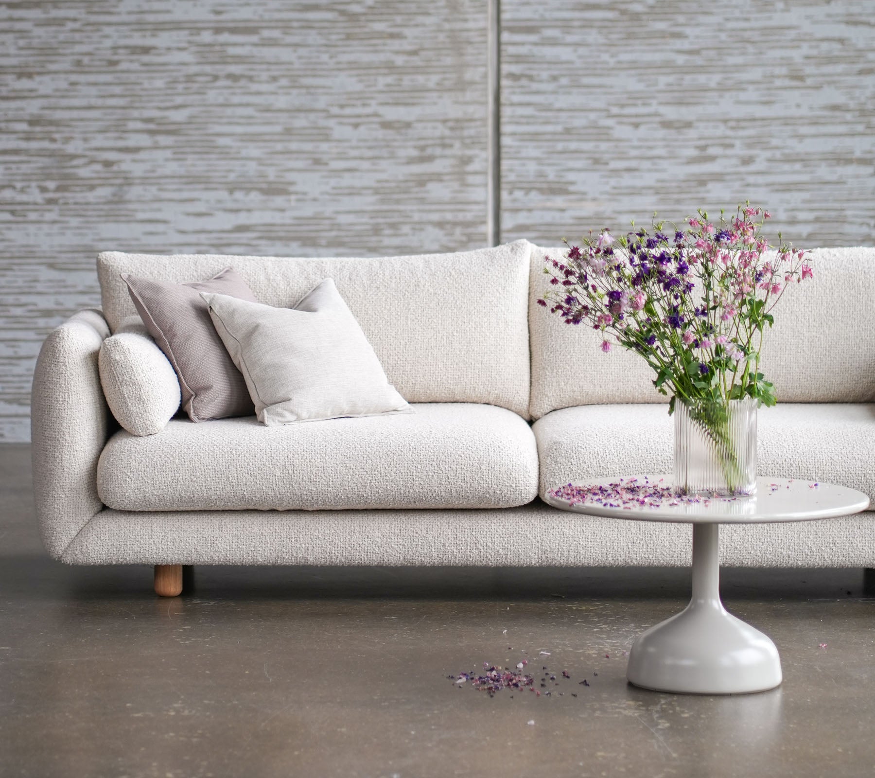 Lysfarvet sofa med teakben, pyntet med puder, suppleret af et rundt bord og en vase med friske blomster.