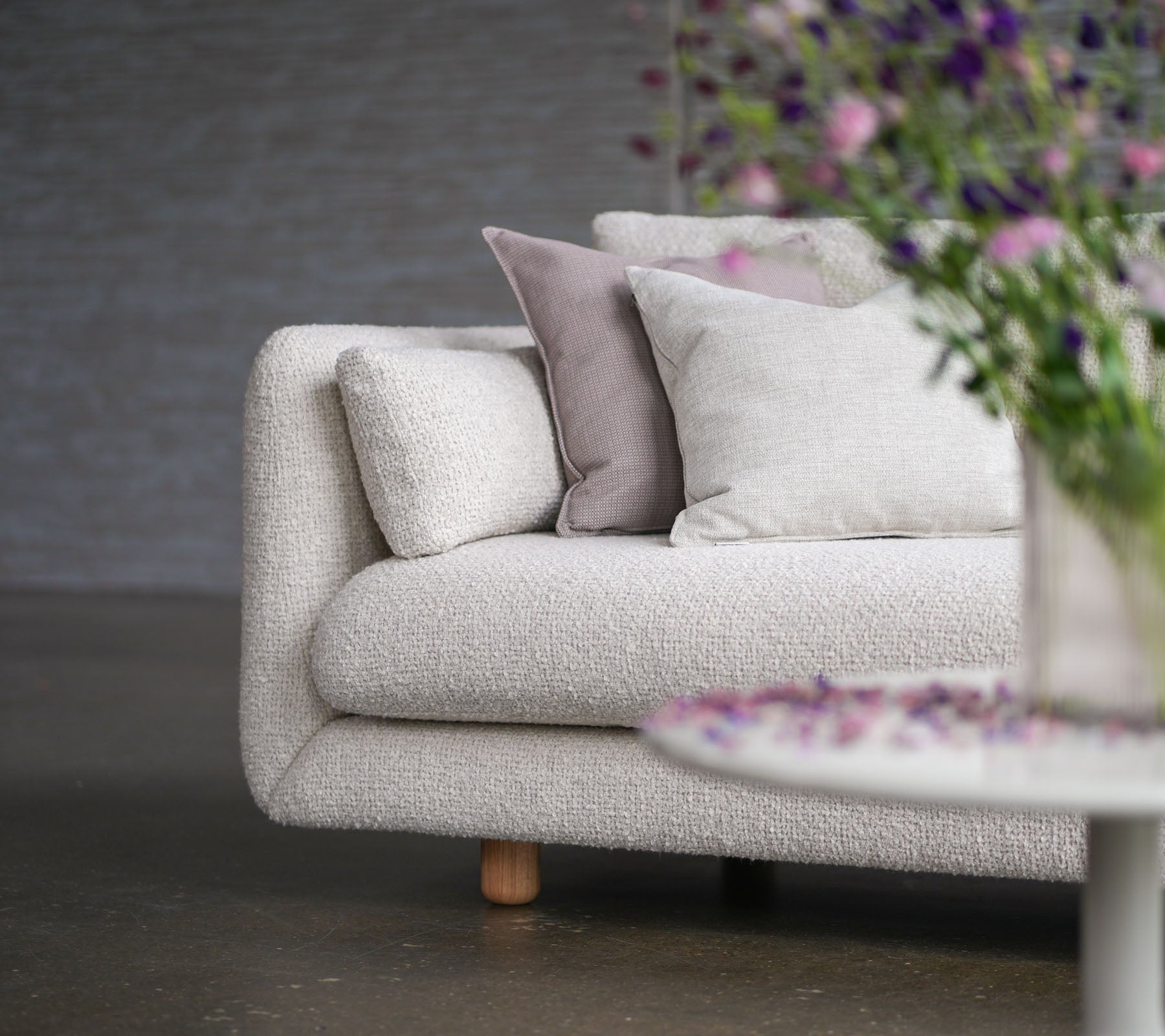 Blød sofa med sandfarvet betræk, dekorative puder, teaktræ ben og en vase med blomster, der viser Cane-line stil og elegance.