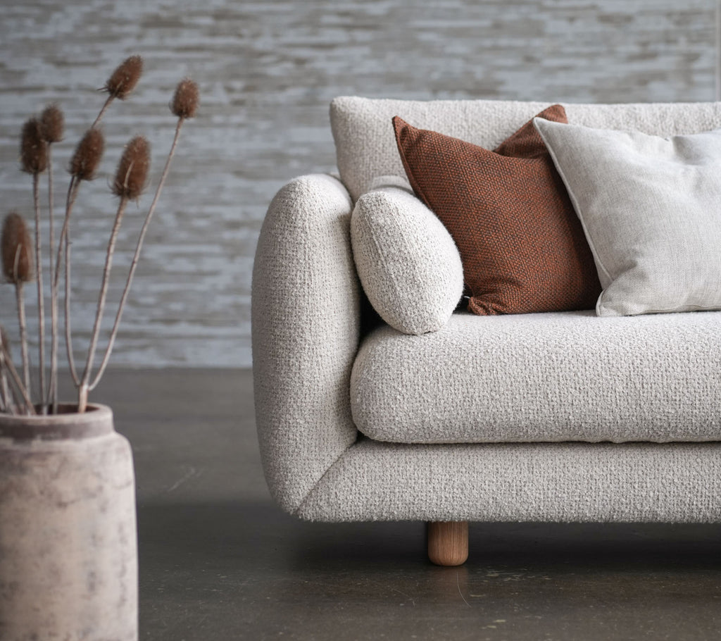 Hyggelig sofa med blomsterbetræk, teaktræben og løftende puder, ledsaget af dekorativ vase og tørrede blomster. Elegant og indbydende design.