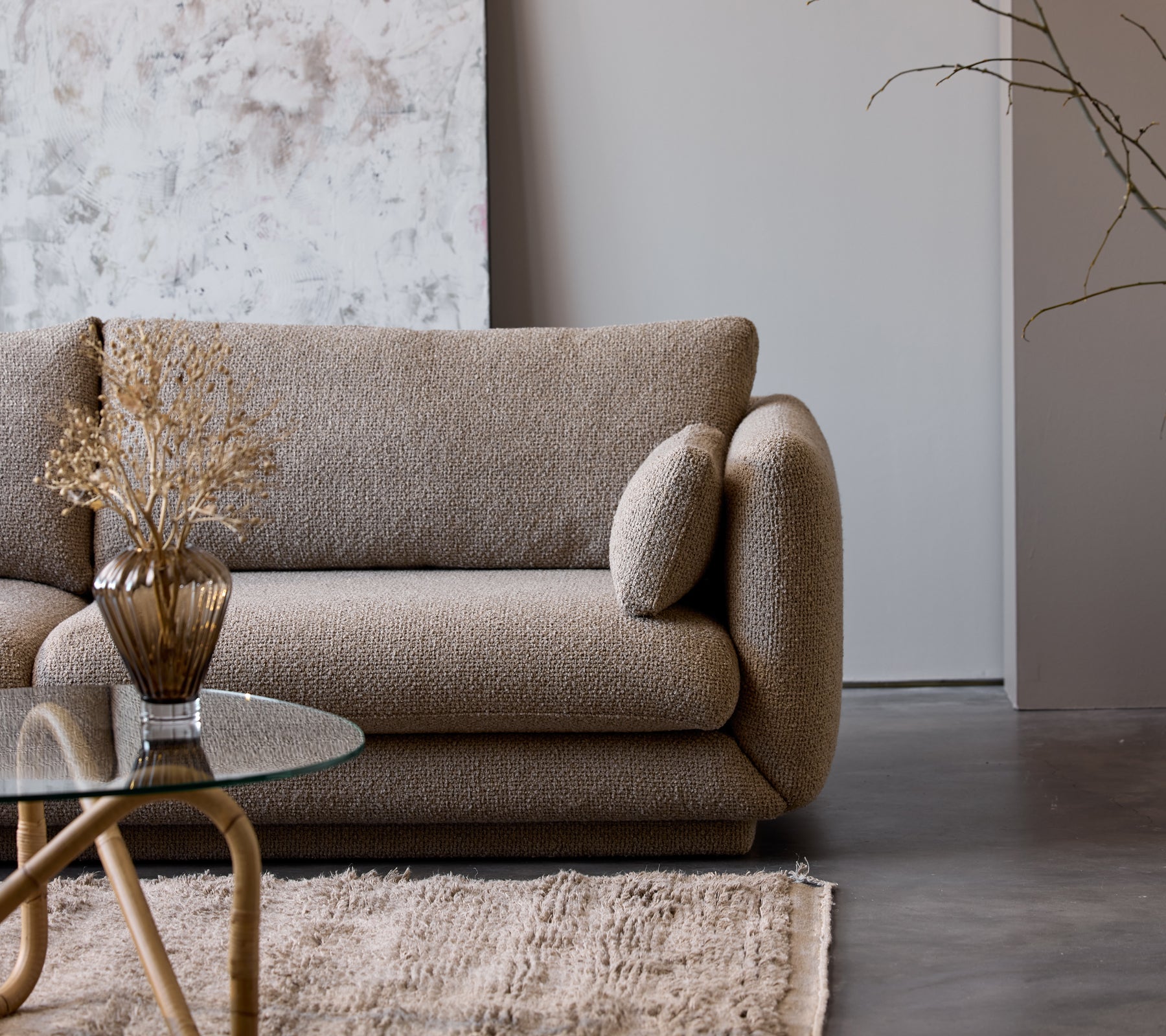 Et taupe farvet sofabord med en glasplade står ved siden af en struktureret beige sofa udsmykket med en dekoreret vase og tørrede blomster.