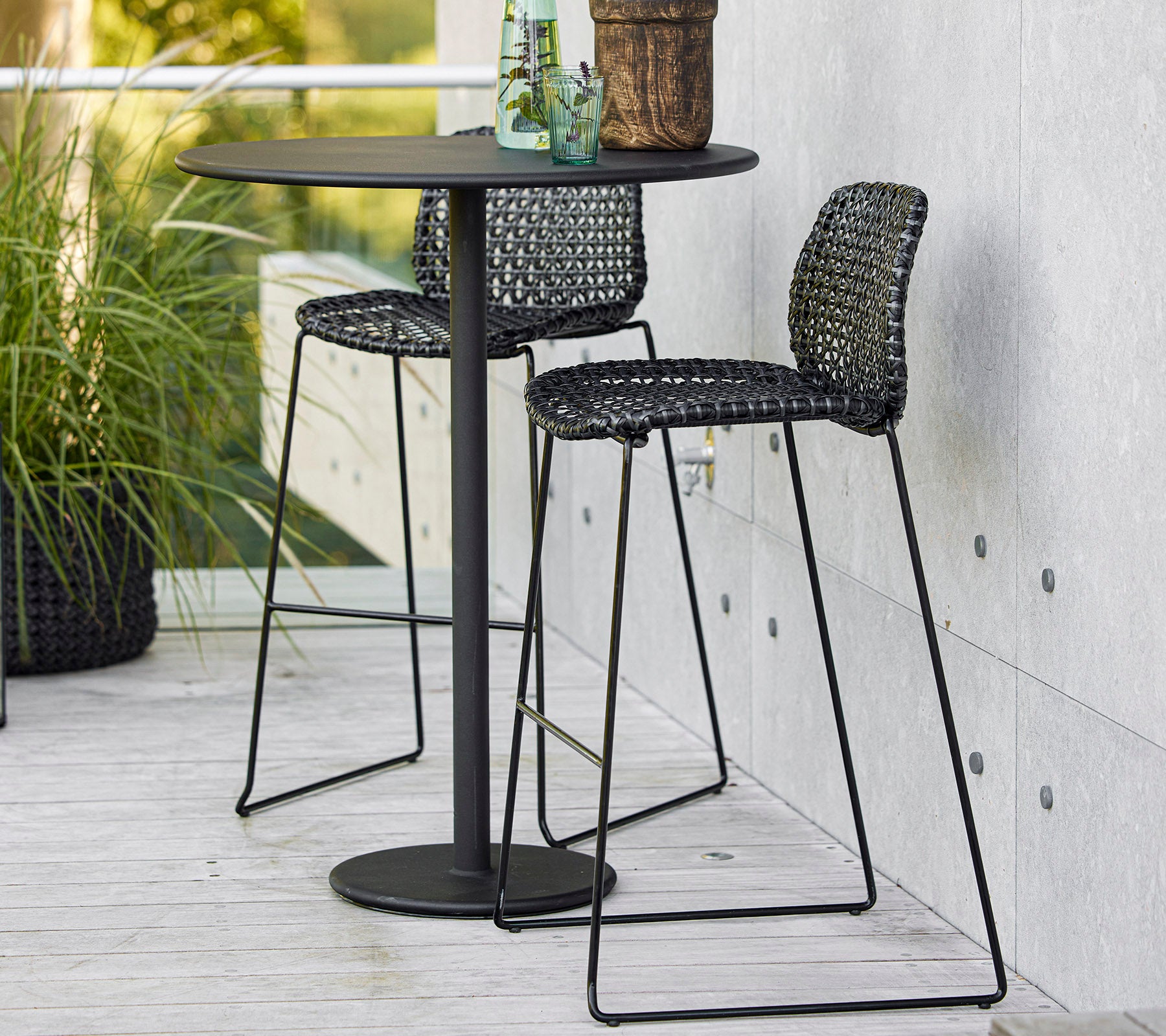Elegant sort bardisk med to vævede stole og en potteplante, perfekt til afslappet spisning eller afslapning.