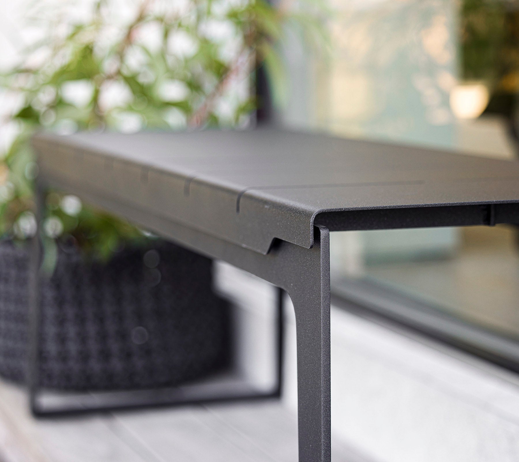 Slank sort bord med et minimalistisk design, der har rene linjer og en robust ramme, suppleret af grønne planter i baggrunden.