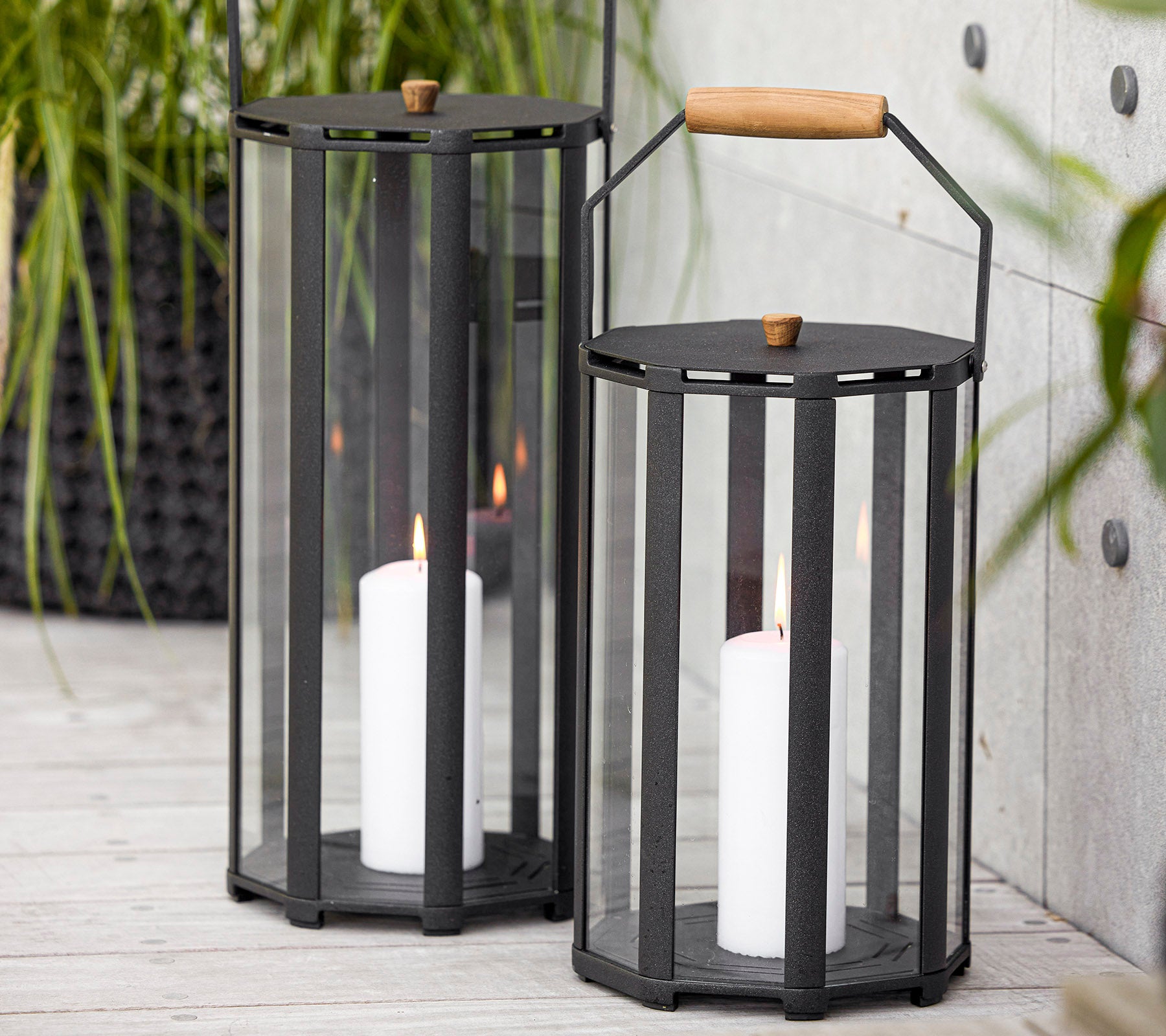 To stilfulde lanterner med et moderne design, der har glaspaneler og et træhåndtag, der holder hvide stearinlys indeni.