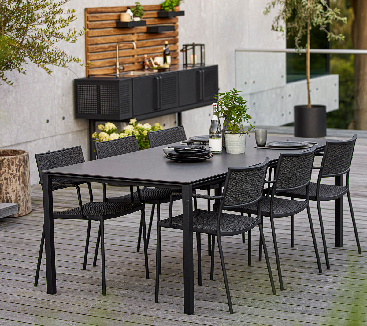 Et moderne dining sæt med et stort rektangulært bord og seks sorte vævede stole, accentueret af dekorative elementer og grønne planter.