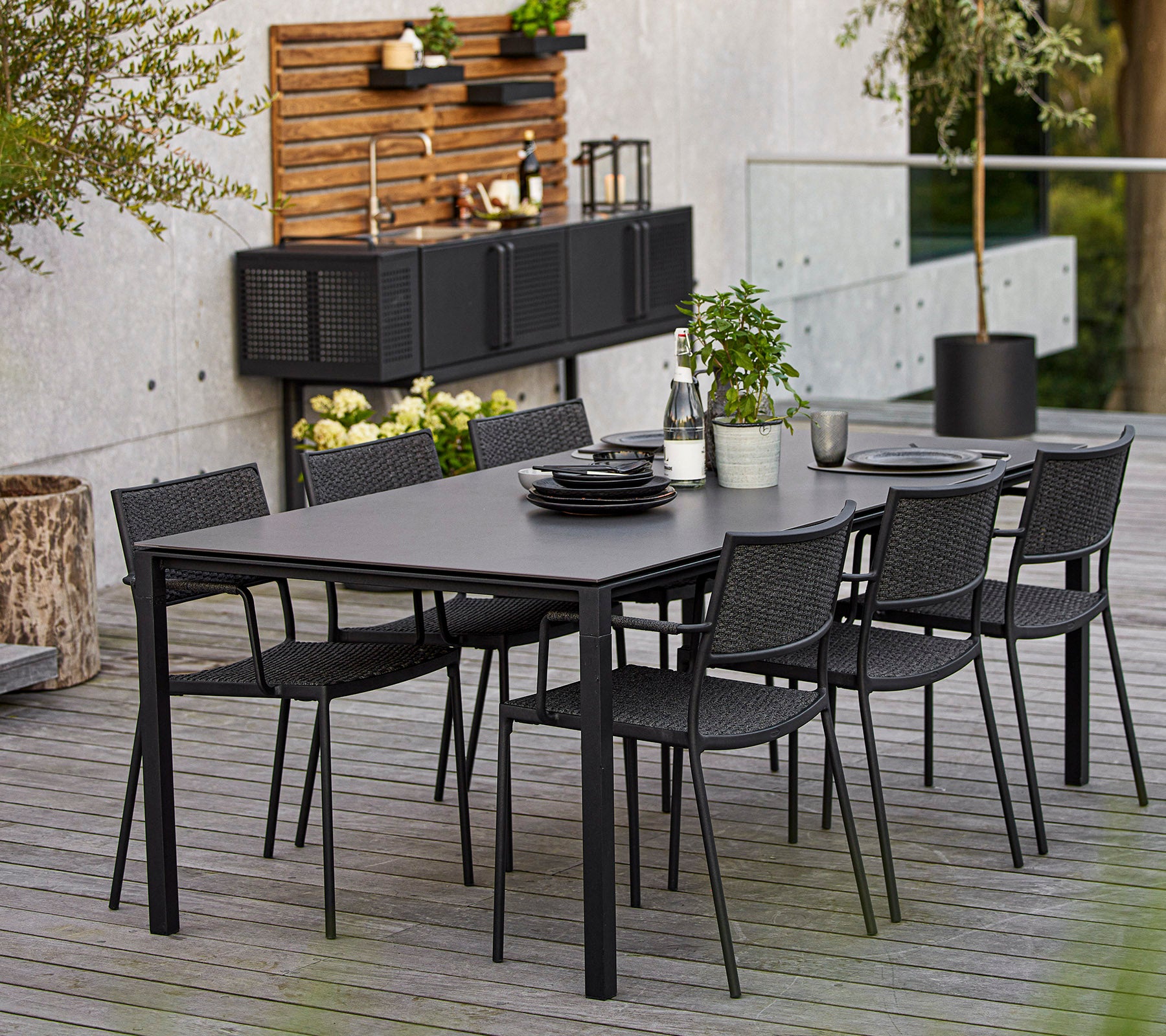 Et moderne dining sæt med et stort rektangulært bord og seks sorte vævede stole, accentueret af dekorative elementer og grønne planter.