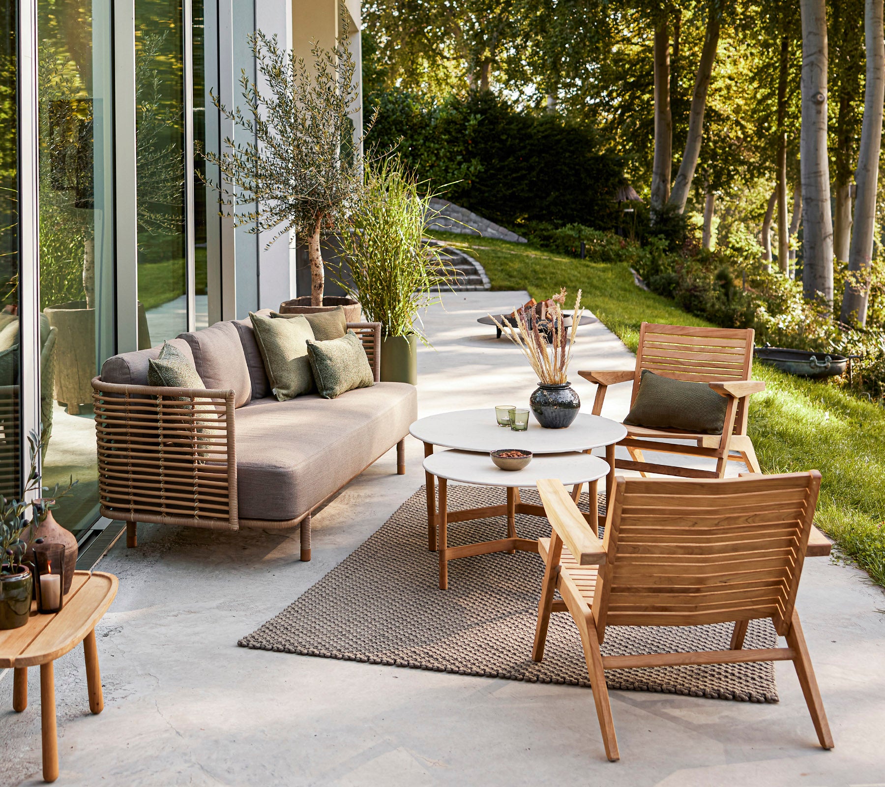 Elegant udendørs siddeområde med en moderne sofa, træstole, et rundt bord og dekorative planter sat mod en fredelig baggrund.