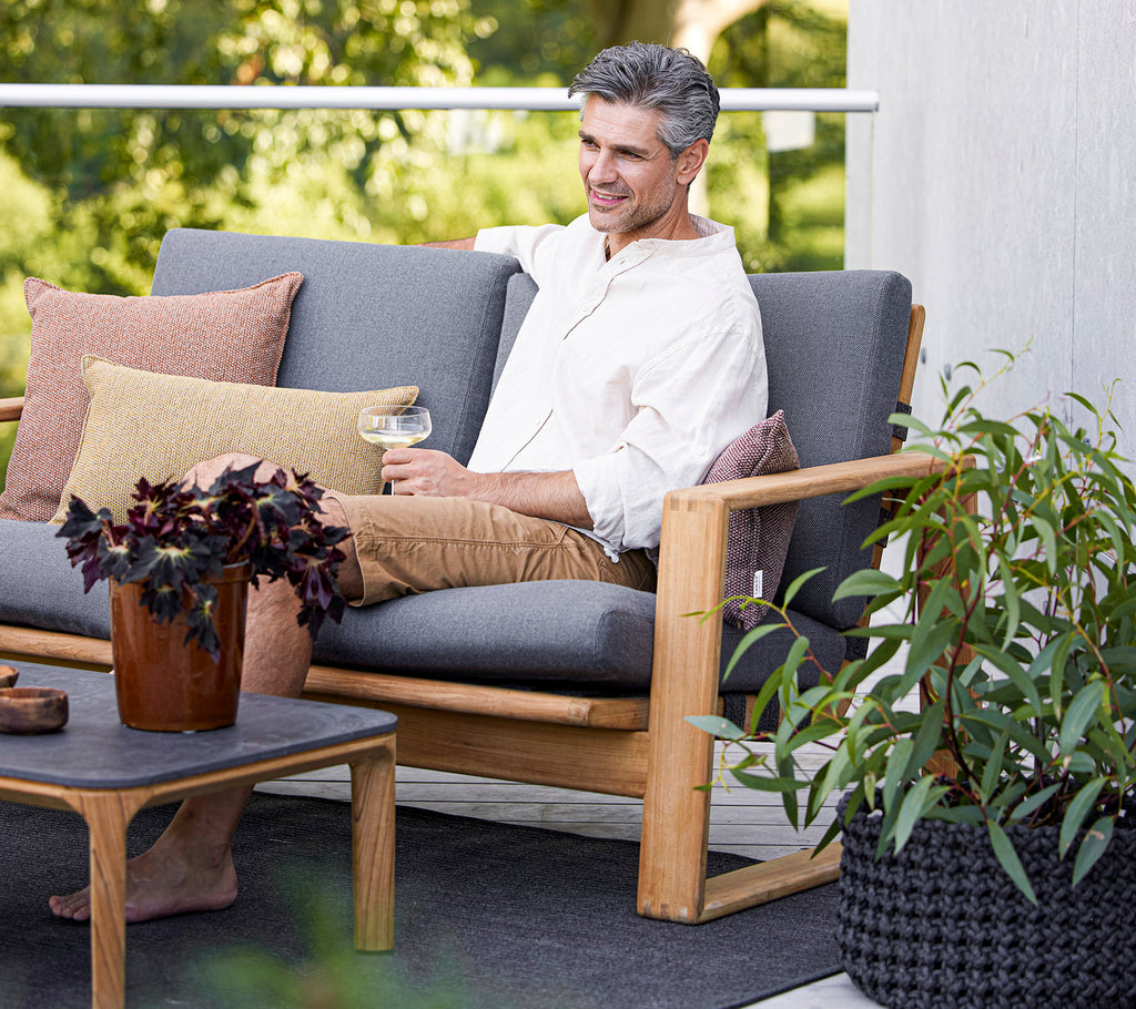 En afslappet mand sidder på en stilfuld sofa med puder, nyder en drink ved siden af en potteplante og et kaffebord.