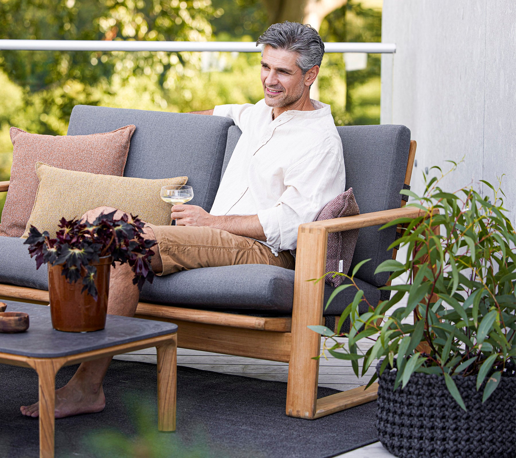 En mand nyder en drink, mens han sidder på en komfortabel sofa, omgivet af planter og dekorative puder.