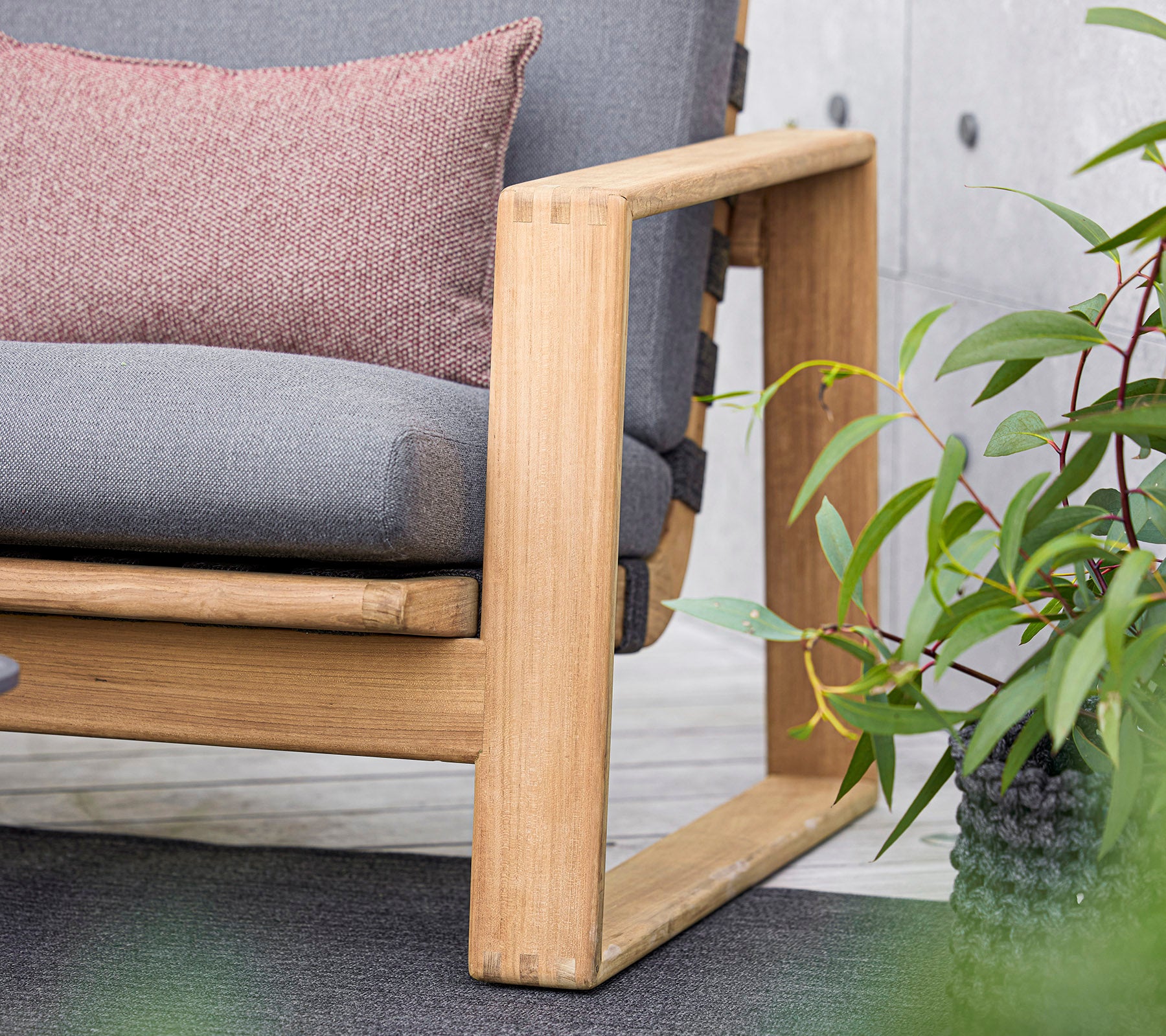 en moderne sofa med en træramme, der har grå puder og en dekorativ pude, suppleret af frodige grønne planter i nærheden.