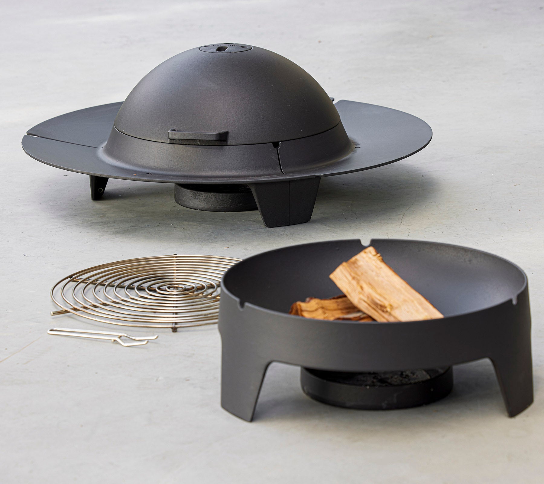 Et moderne bålsted sættet med et slankt, cirkulært design med et kuppellåg, ledsaget af et grillrack og træstykker.
