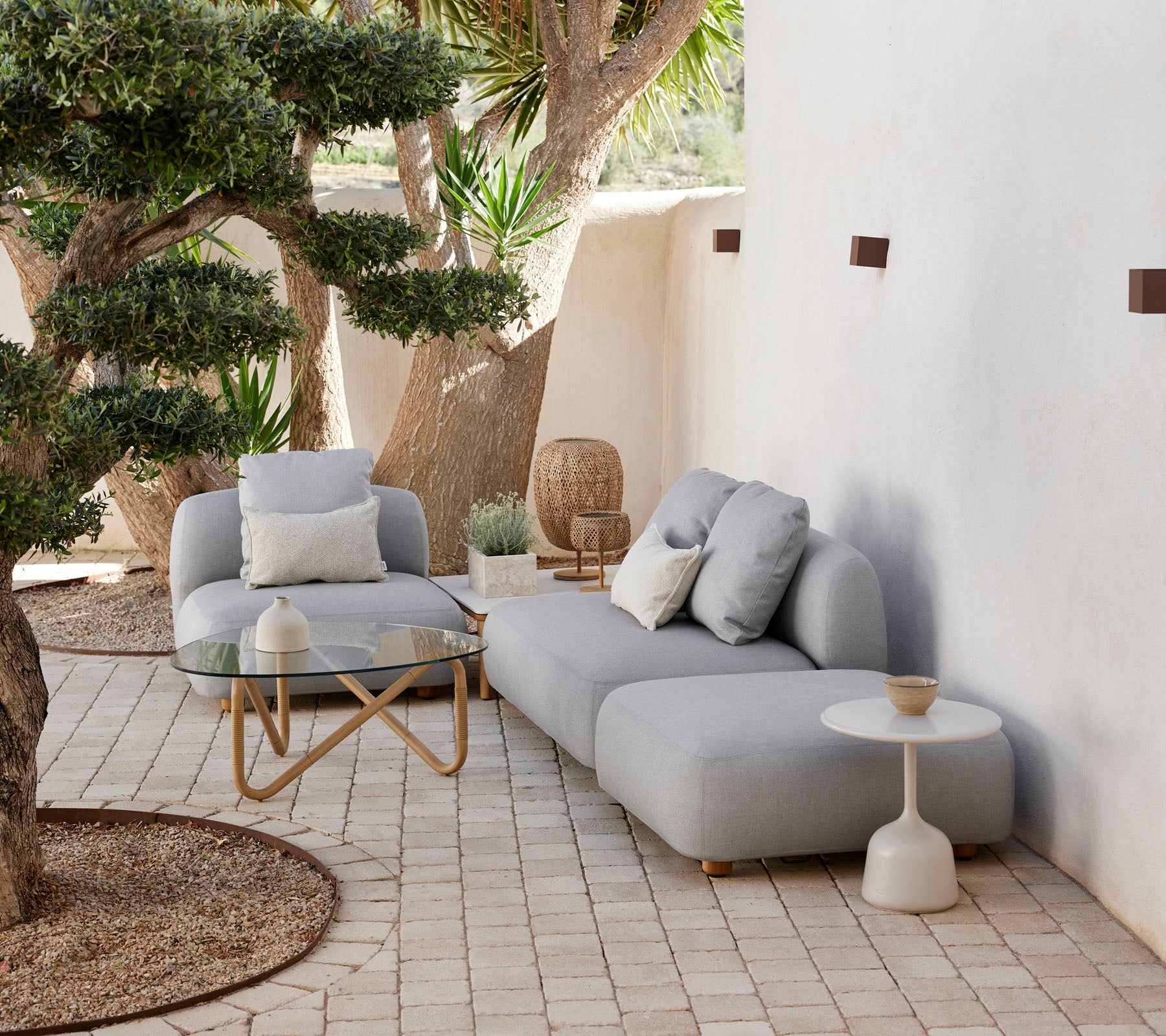 Moderne udendørs siddearrangement med en hyggelig sofa, accentstol og stilfuldt sidebord, omkranset af grønne planter og dekorative elementer.