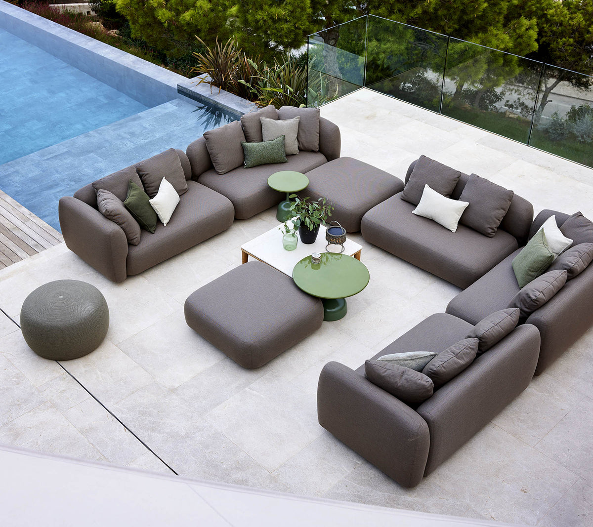 Et moderne loungemiljø med et sektionelt sofaarrangement, rund ottoman og grønne accentborde, omgivet af grønne planter og en fredfyldt pool.