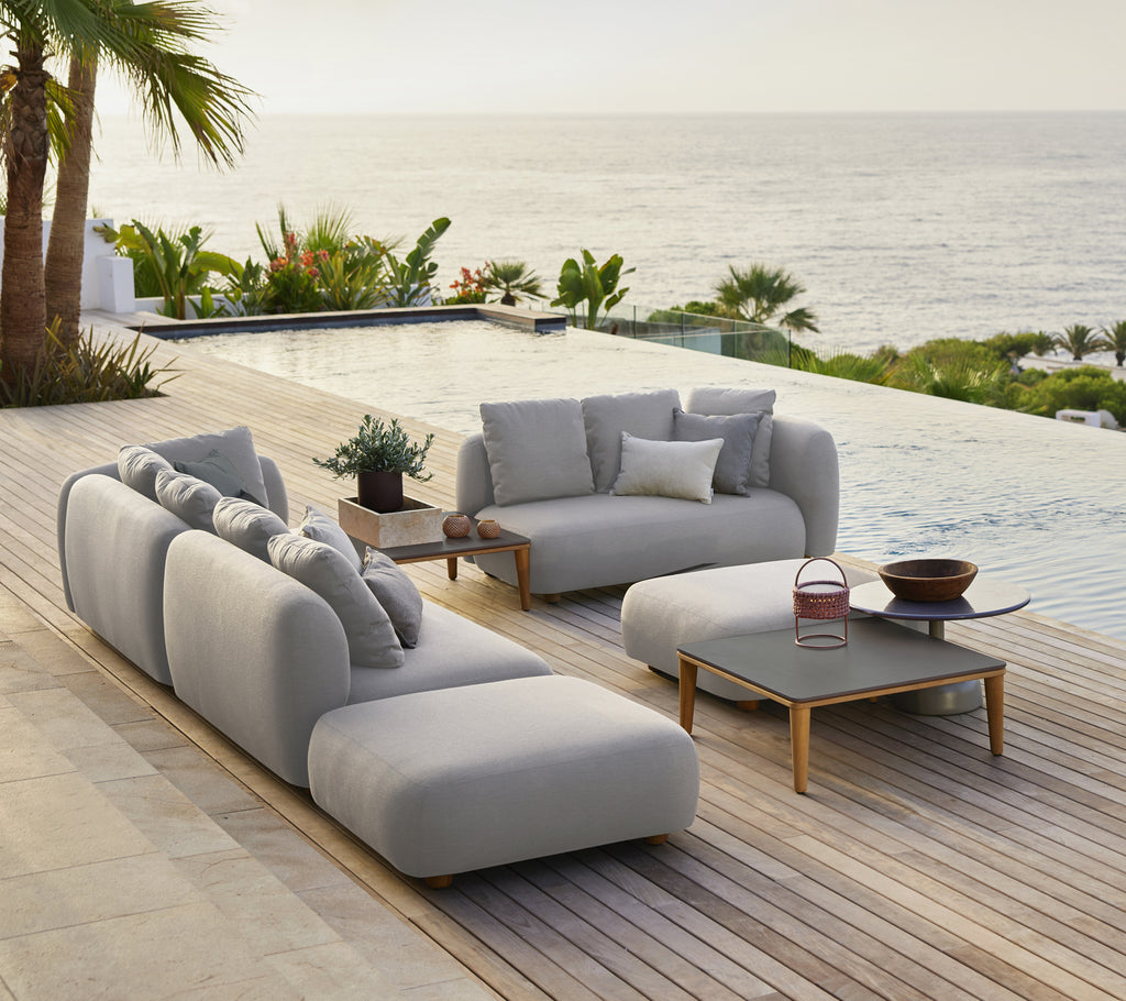 Stilfuld grå sofa med puder og et sort kaffebord i en moderne udendørs lounge ved havet.