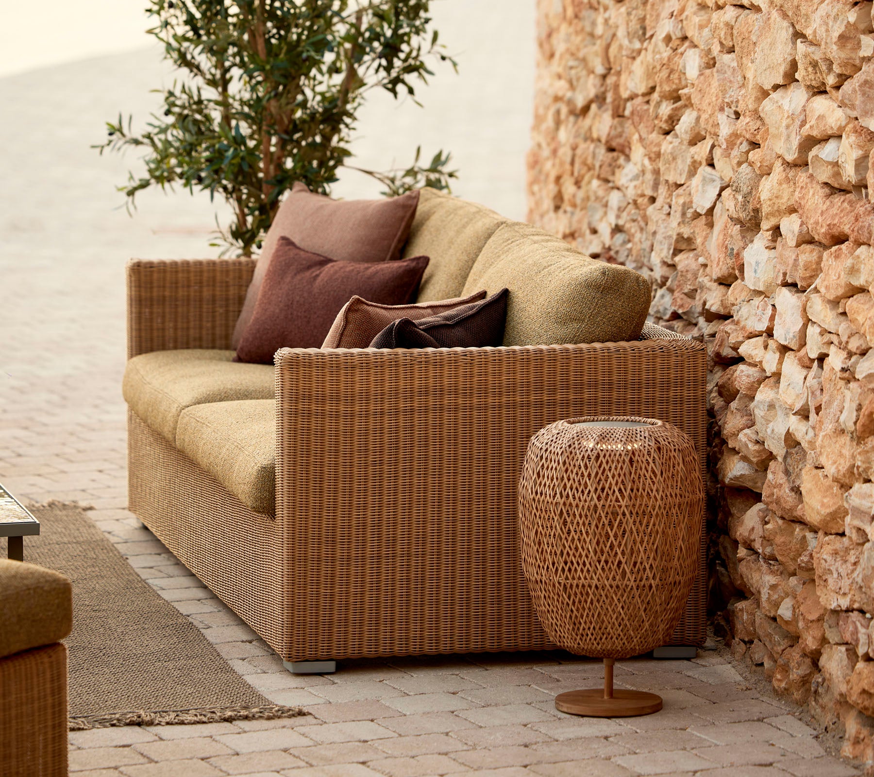 En hyggelig rattansofa med puder, suppleret af et stilfuldt vævet sidebord, der står foran en stenvæg og frodige planter.