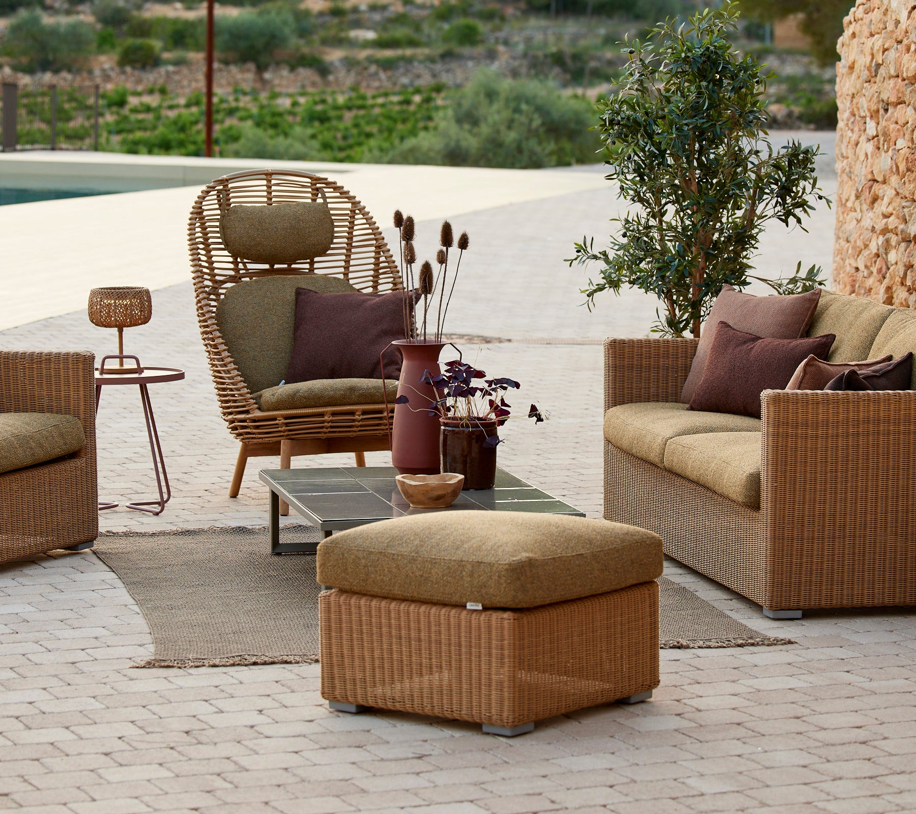 Lounge siddeområde med rattan stol, beige sofa og sidebord med en lilla vase.