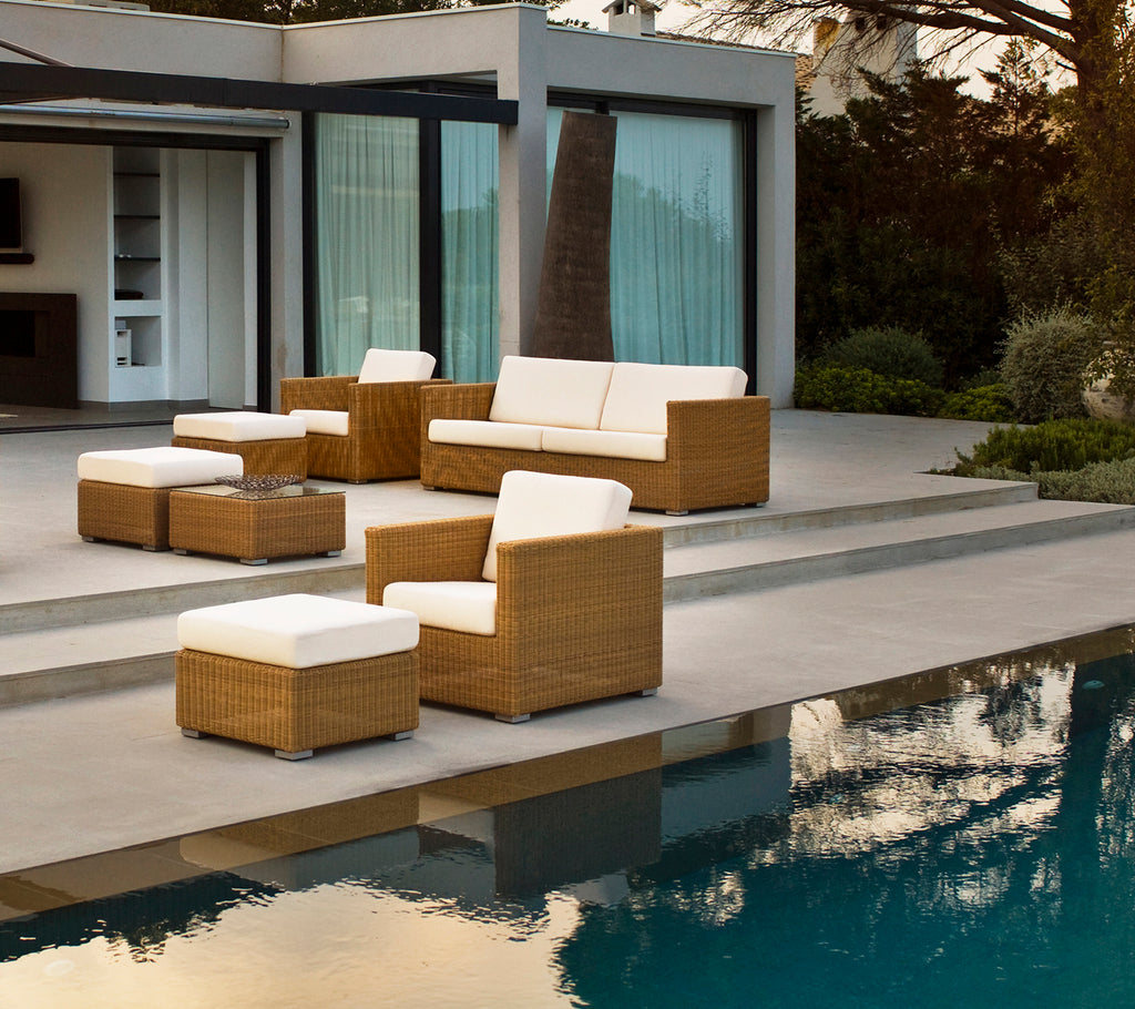 Moderne udendørs loungeområde med brun sofa, stole og puder ved en pool.