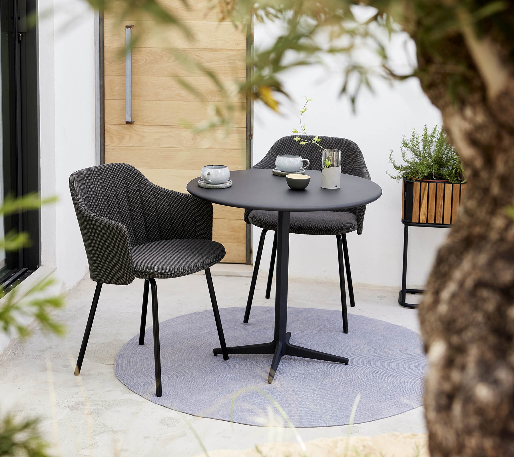 Stilfuldt valg af stol og rundt café bord sæt, ledsaget af en kop og underkop, omgivet af grønne planter.