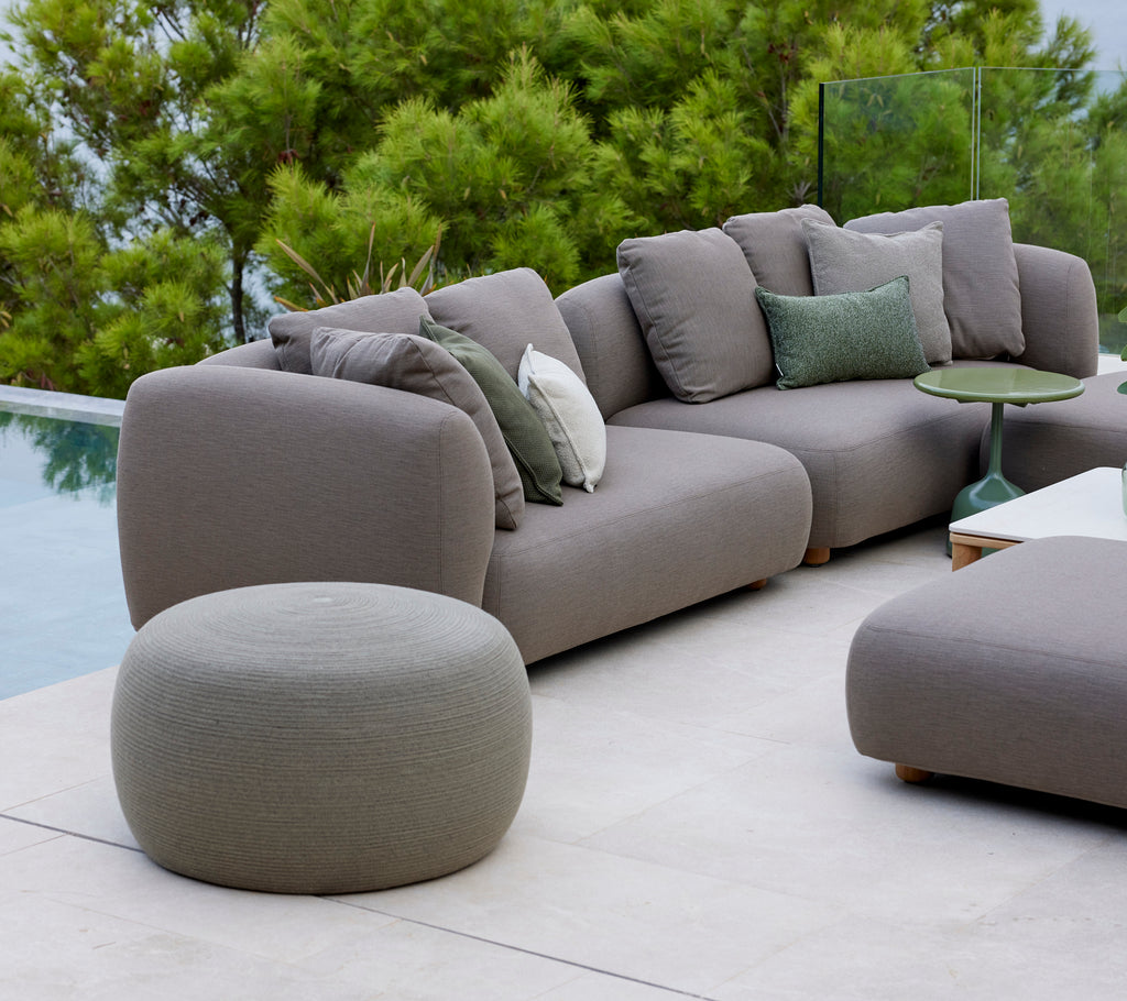 Stor taupe-farvet cirkelfodskammel ved siden af en moderne sofa med grønne puder, omfavnet af frodig grønhed og en fredfyldt atmosfære.