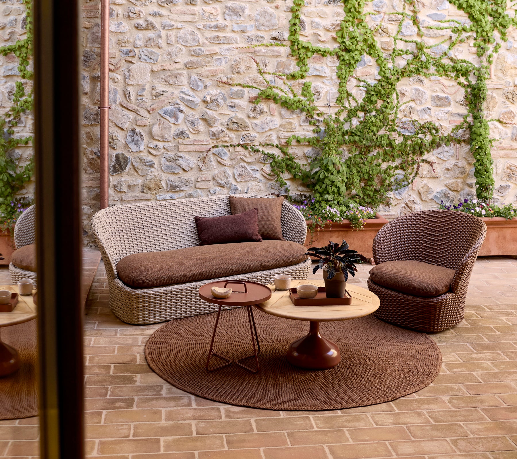 Hyggeligt udendørs loungeområde med en vævet sofa, en stol, et rundt sidebord og grønne planter mod en stenmur.