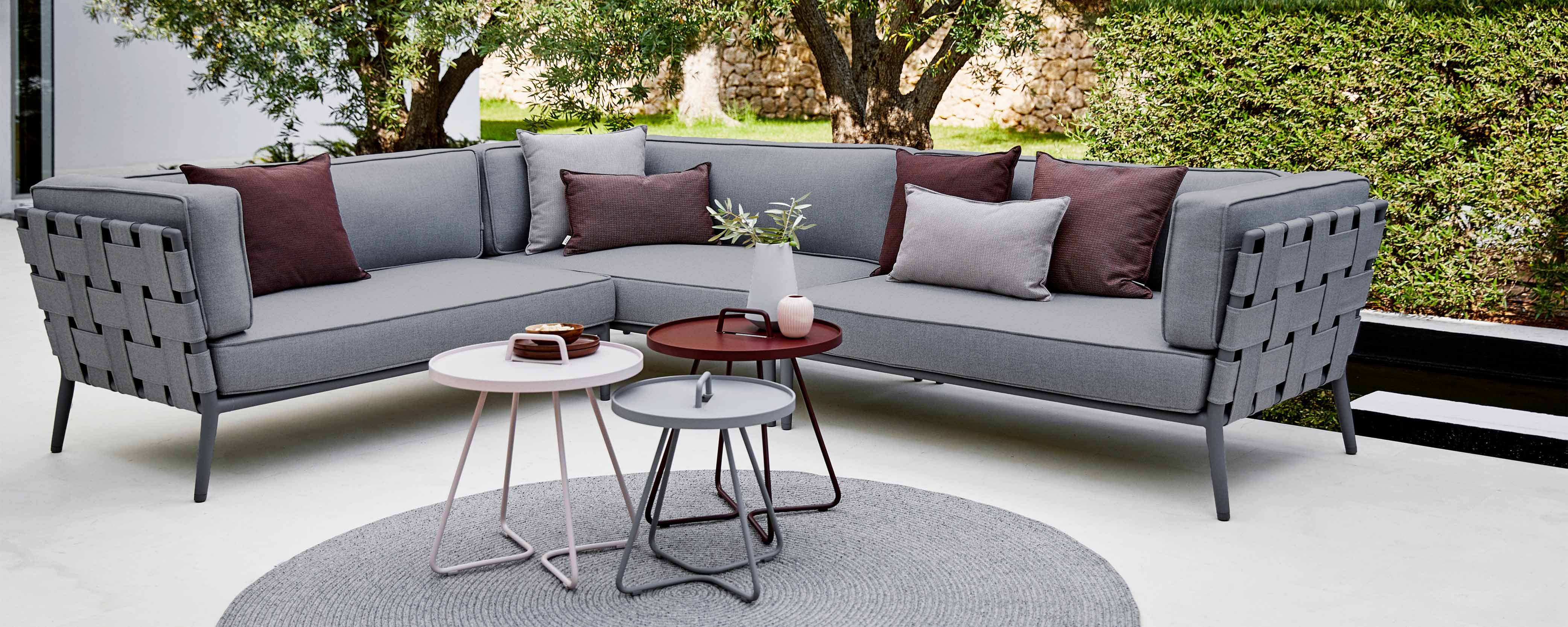 Moderne konisk sektionel sofa i gråt stof med dekorative puder, ledsaget af stilfulde sideborde og et rundt tæppe.