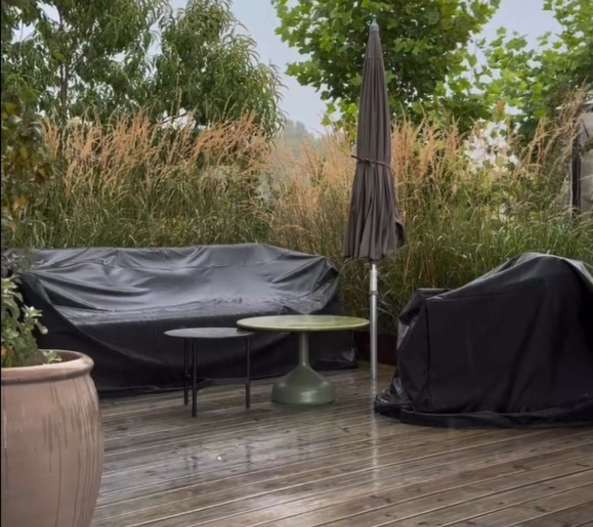 Udendørsområde med sort sofa, grøn bord og sort stol omgivet af grønne planter.