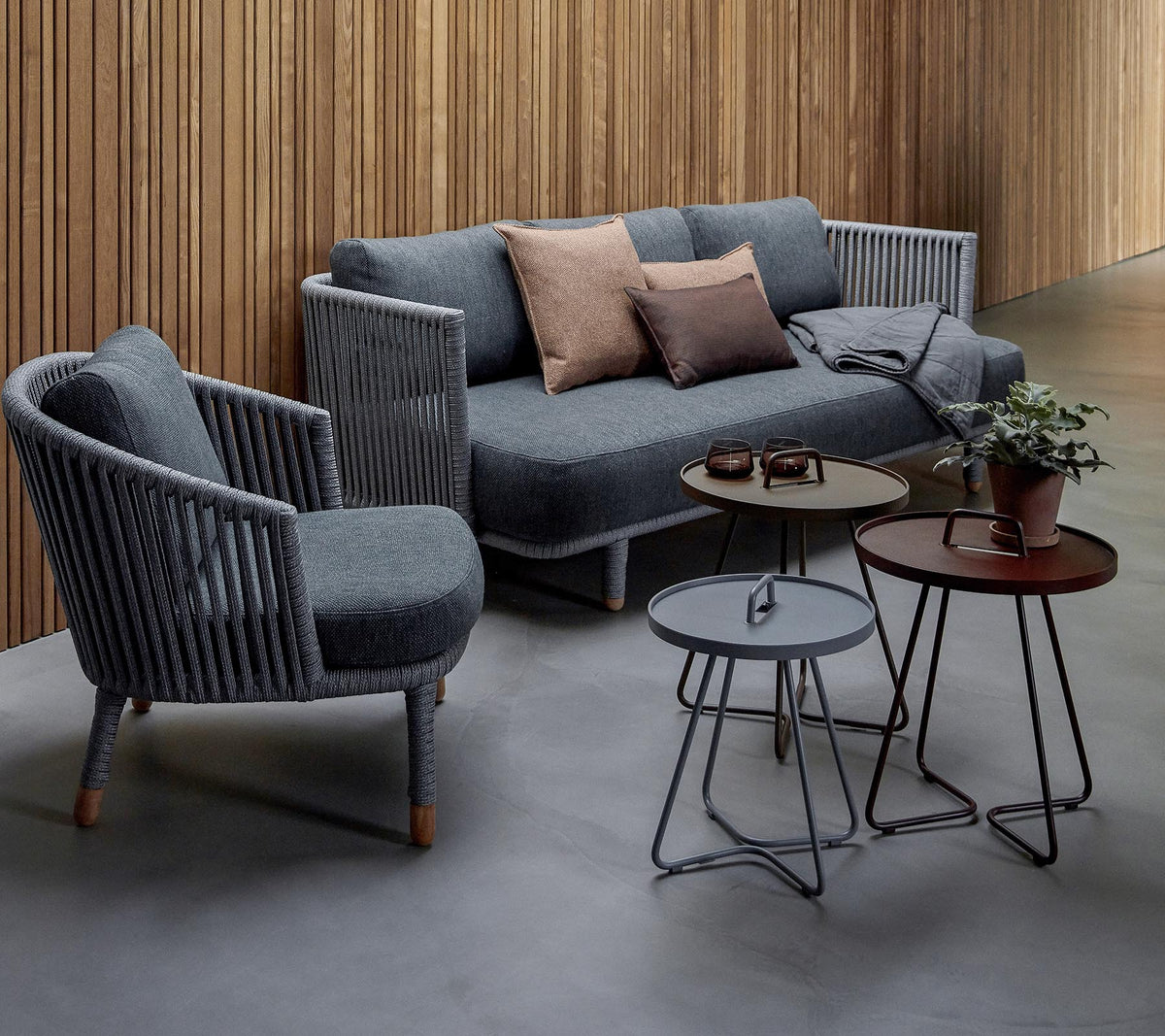 Stilfuld siddearrangement med en grå sofa med puder og to accentstole, ledsaget af tre moderne borde og en potteplante.
