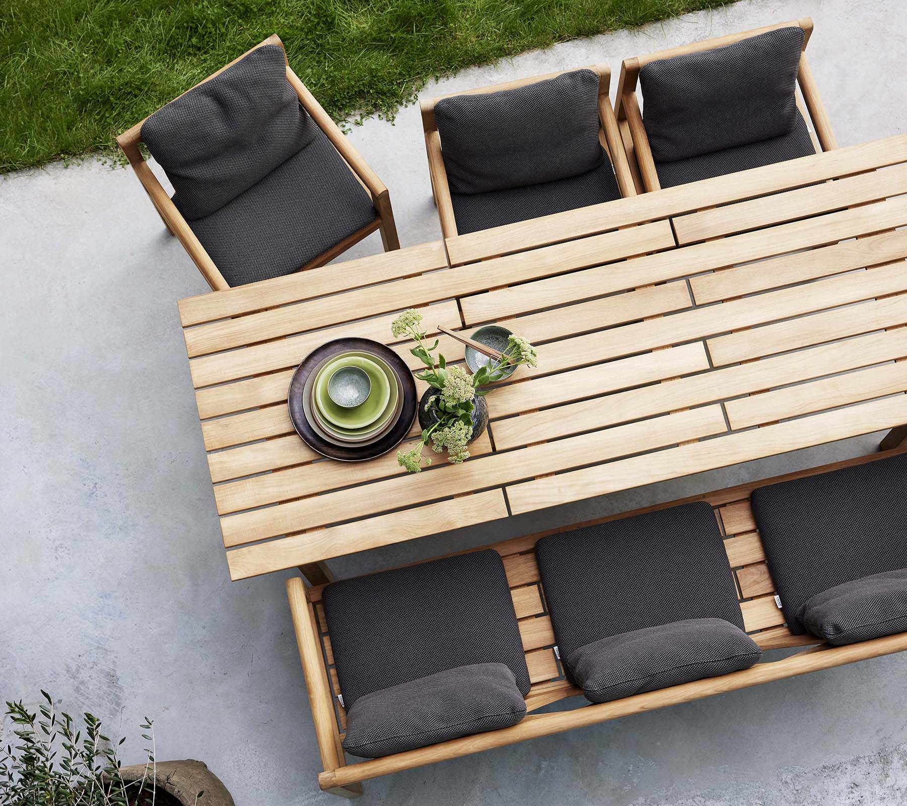 Et træ spisebord med en bænk og stole med mørke puder, accentueret af dekorative tallerkener og grønne planter.