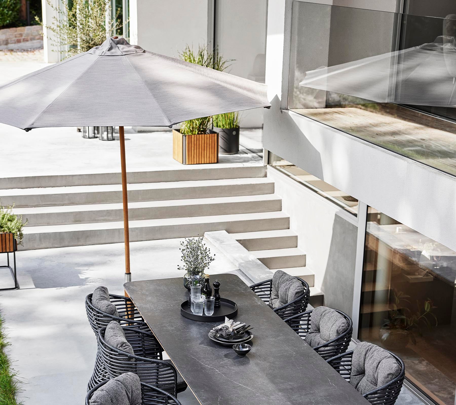 Et stilfuldt udendørs spiseområde med et langt bord, kurvestole og en klassisk parasol til skygge.