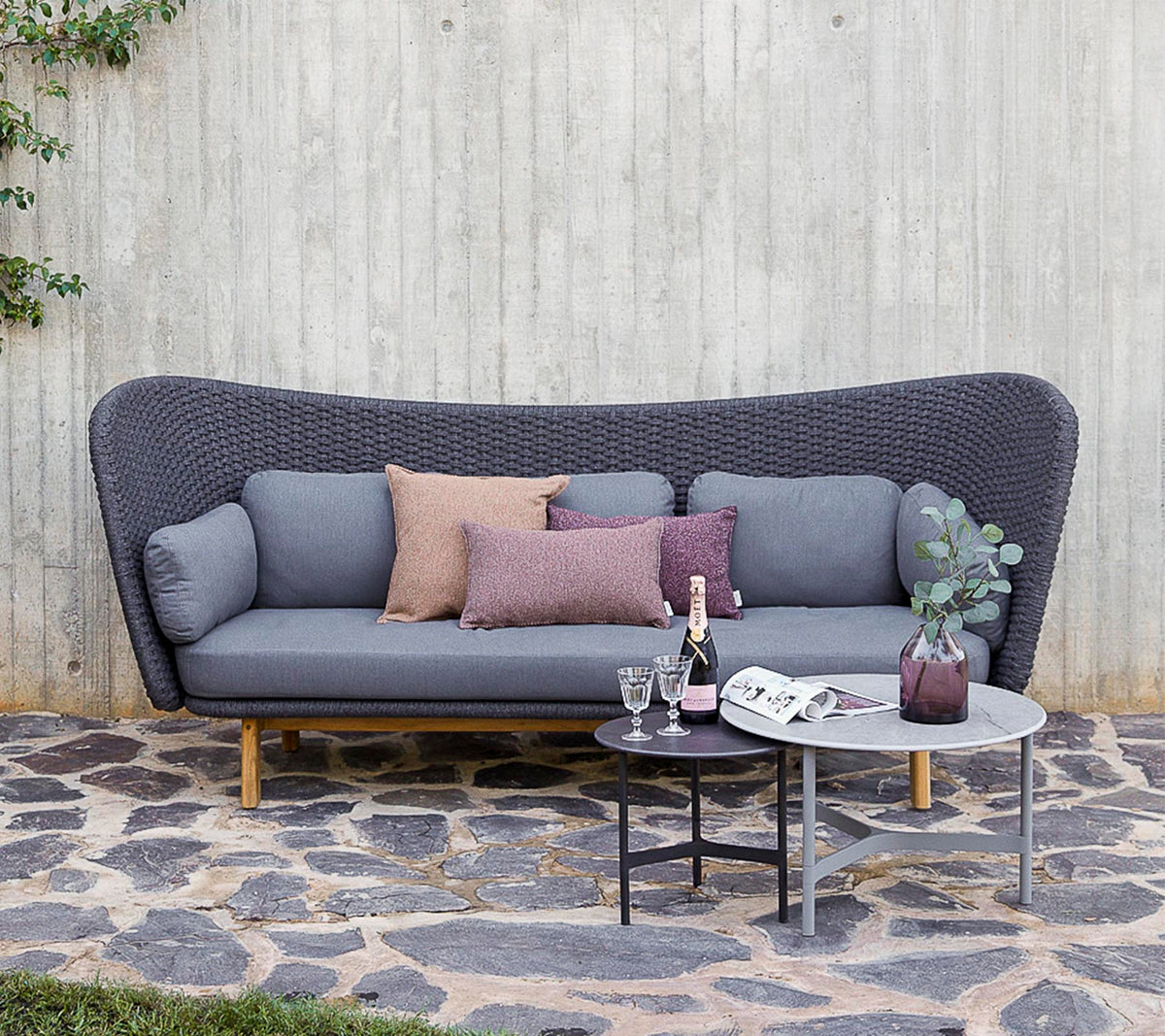 En moderne sofa i grå med dekorative puder, ledsaget af et lille bord med drikkevarer og en vase, sat mod en struktureret væg.