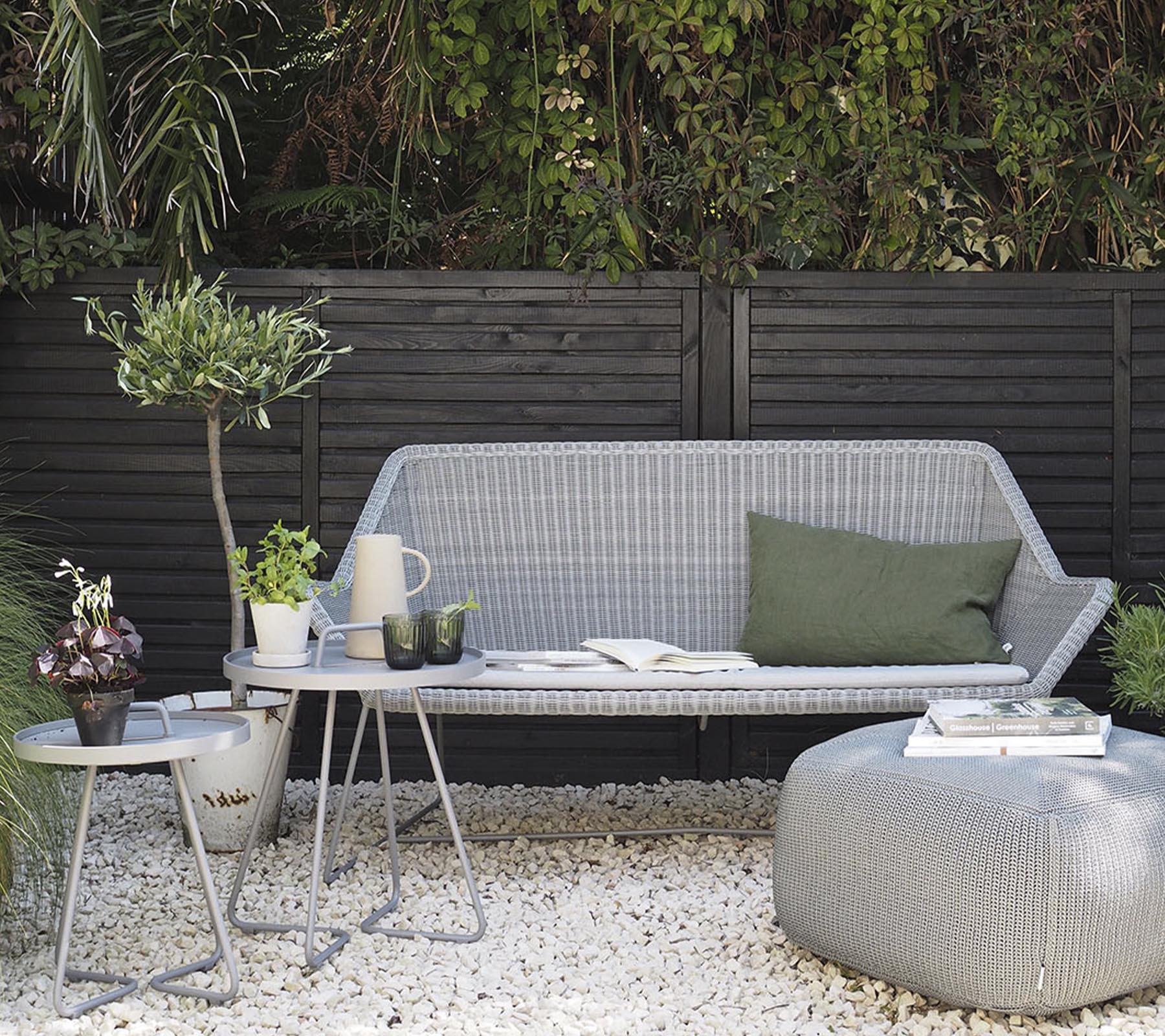Minimalistisk udendørs møbelarrangement med en stilfuld sofa, sideborde og dekorative planter på en grusoverflade.