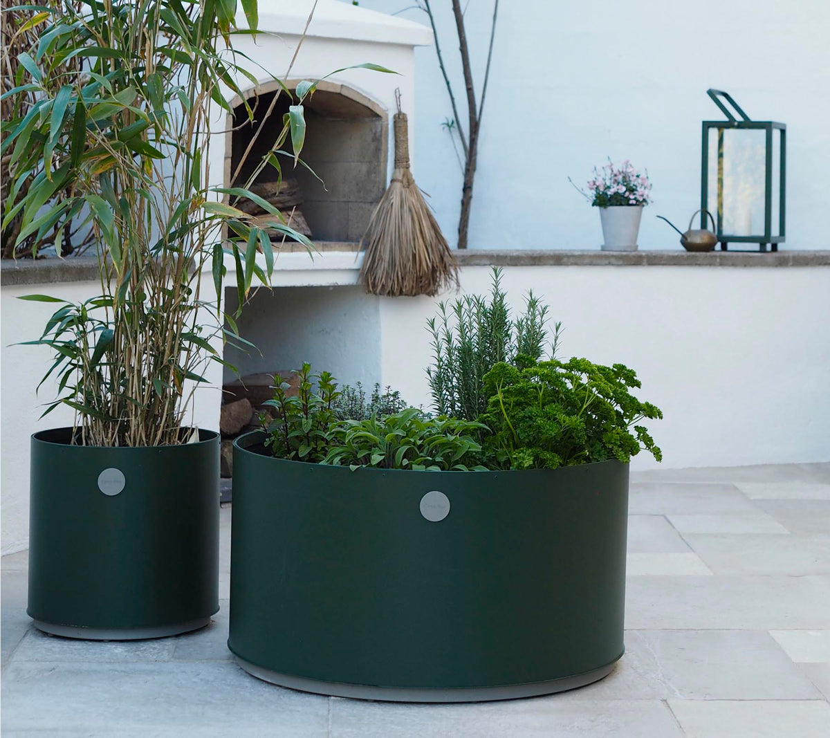 To runde plantekasser i mørkegrøn, den ene med høje planter og den anden fyldt med forskellige urter, står på en lys stensurface.