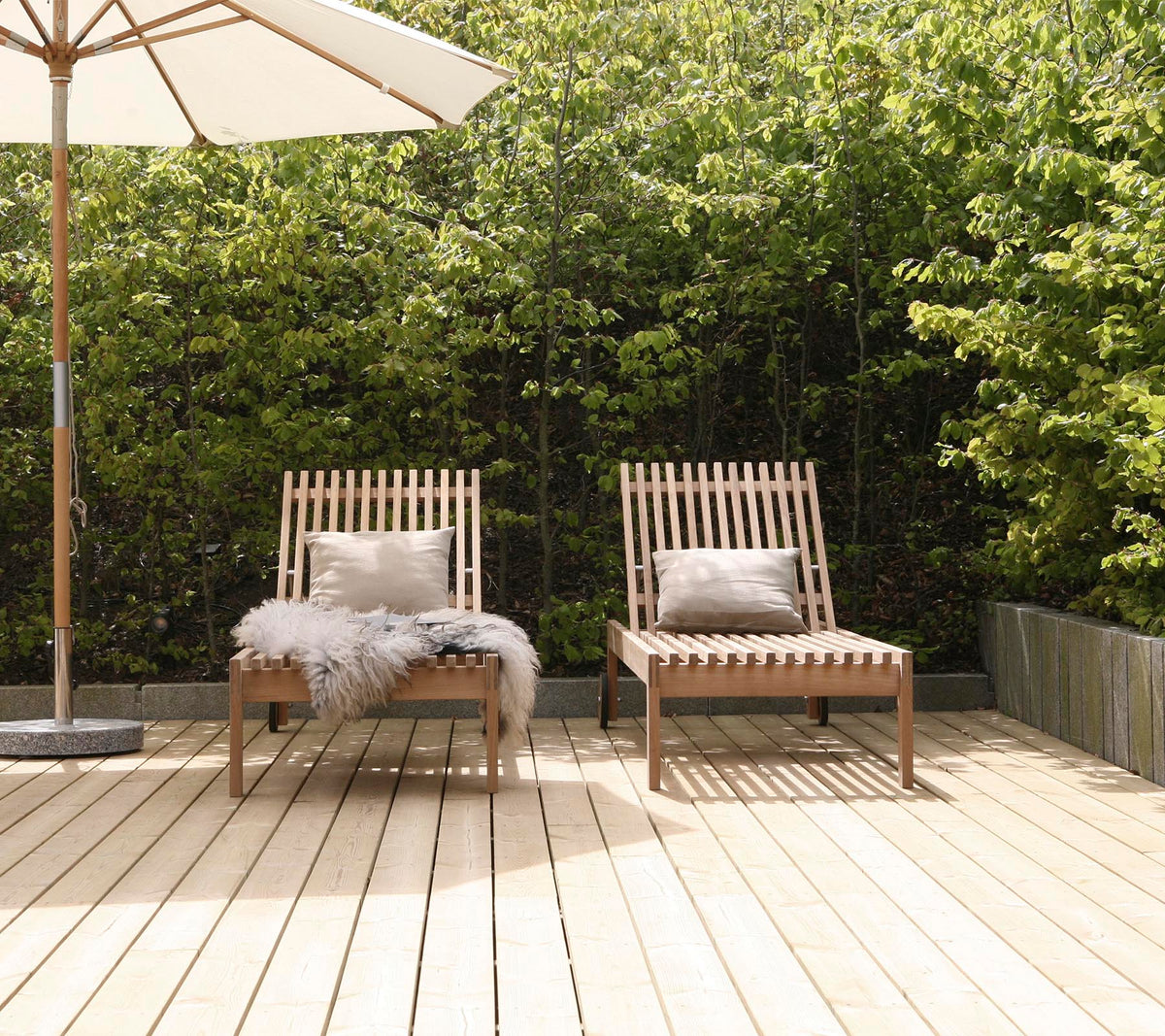To stilige træloungestole med puder, placeret på en træterrasse, omgivet af grønne planter og en stor parasol i nærheden.