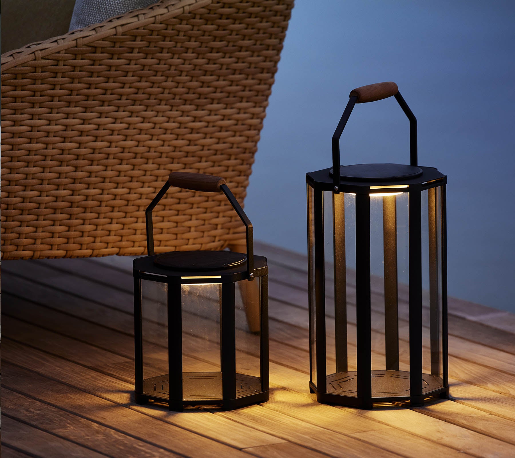 To stilfulde Lightluxe lanterner med glaspaneler og håndtag, der udsender et varmt skær, placeret ved siden af en vævet stol.