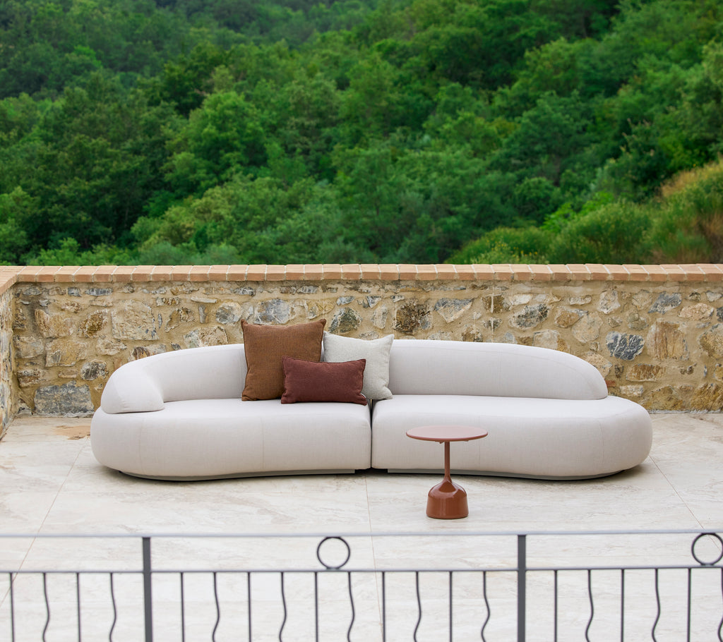 En moderne, buet sofa med neutral polstring og dekorative puder, suppleret med et stilrent sidebord i en naturlig omgivelser.
