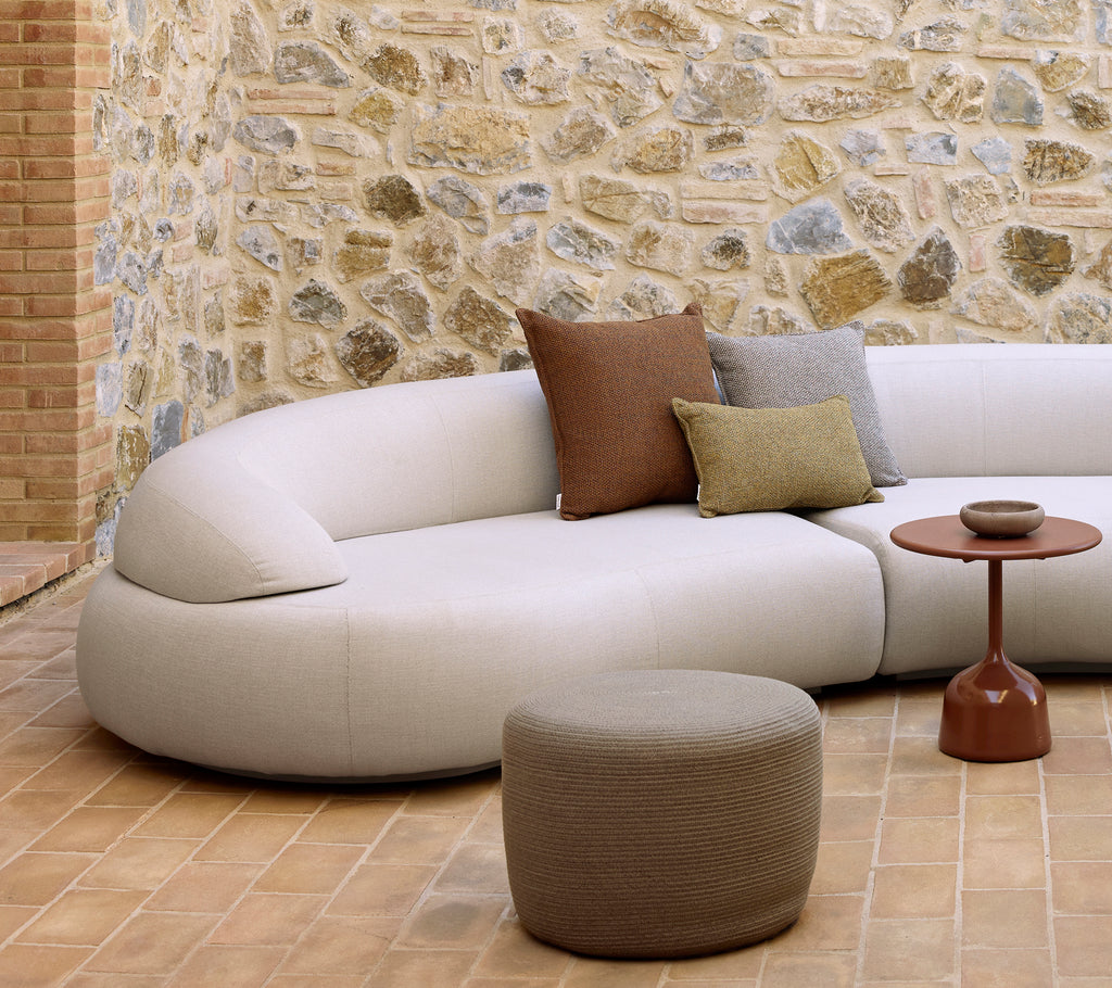 Krum sofa med dekorative puder, en cirkulær fodskammel og et glaseret sidebord på et tekstureret gulv med en stenvæg i baggrunden.