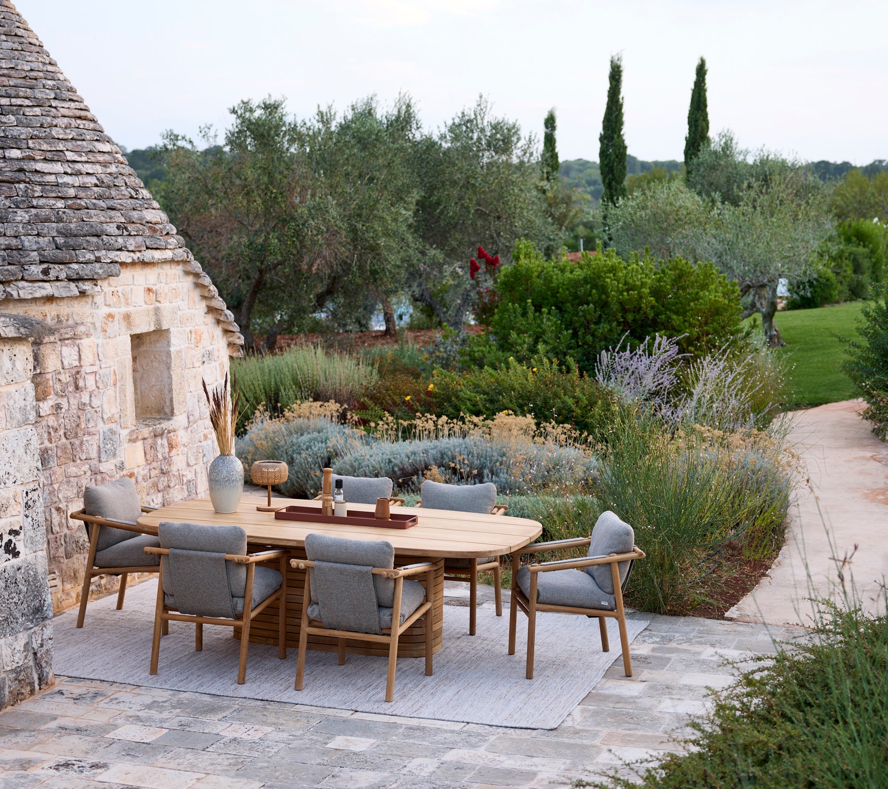 Moderne udendørs spisestue med et rundt bord og grå stole omgivet af grønne planter.