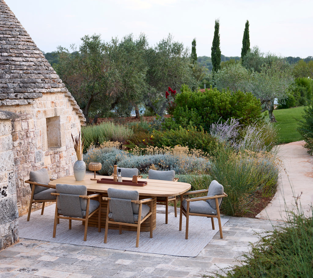 Moderne udendørs spisestue med et rundt bord og grå stole omgivet af grønne planter.